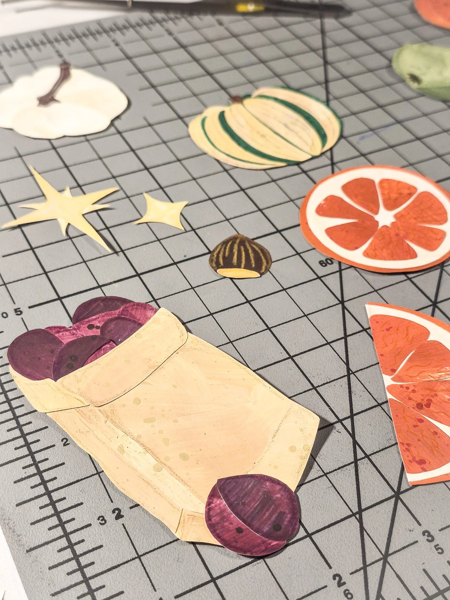 A collage worktable covered with painted paper pieces, citrus slices, squash, mushrooms, and color studies.