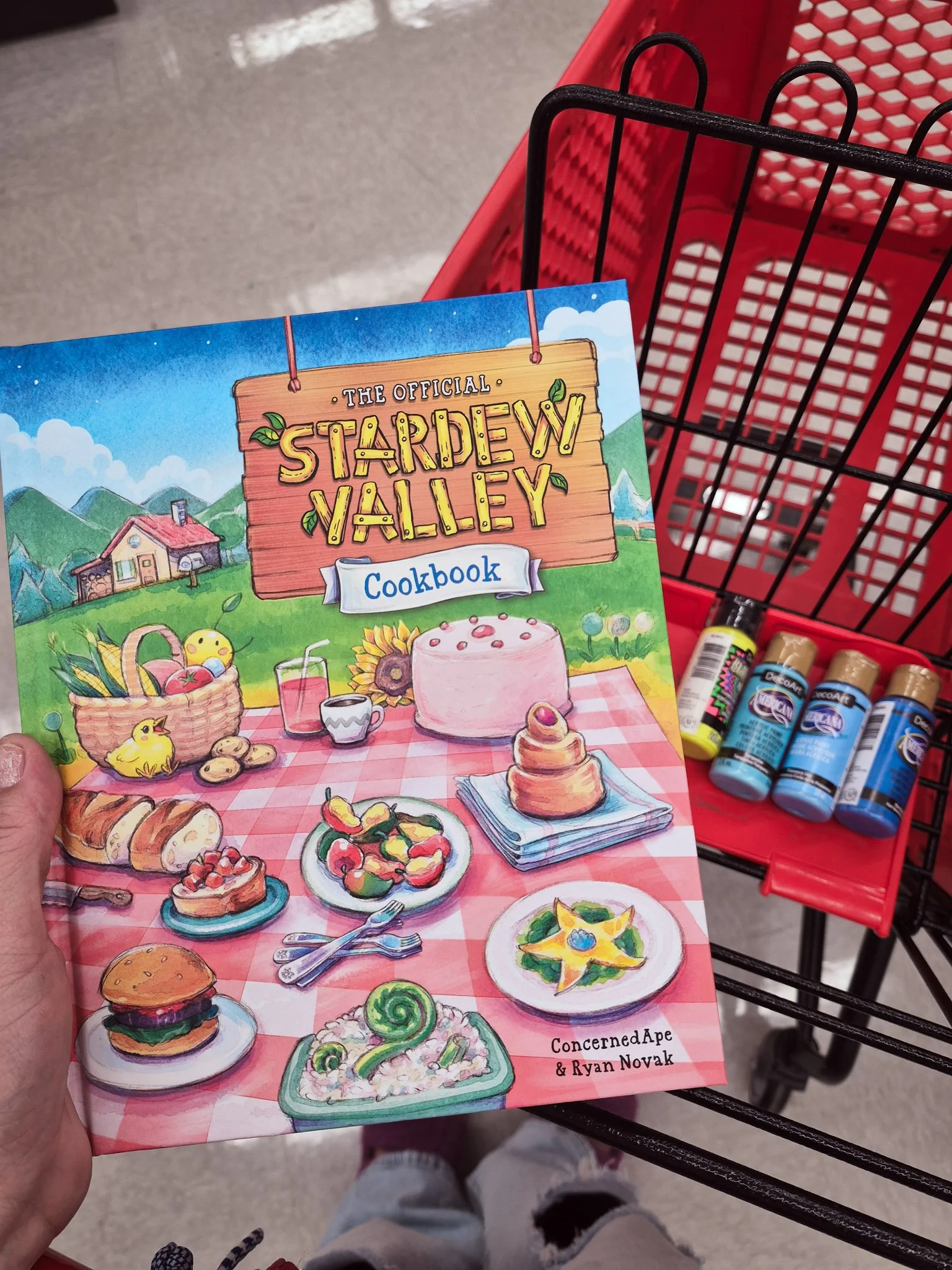 an image of a red Michael's shopping cart with paint tubes in the background while the artist is holding a copy of The Stardew Valley Cookbook
