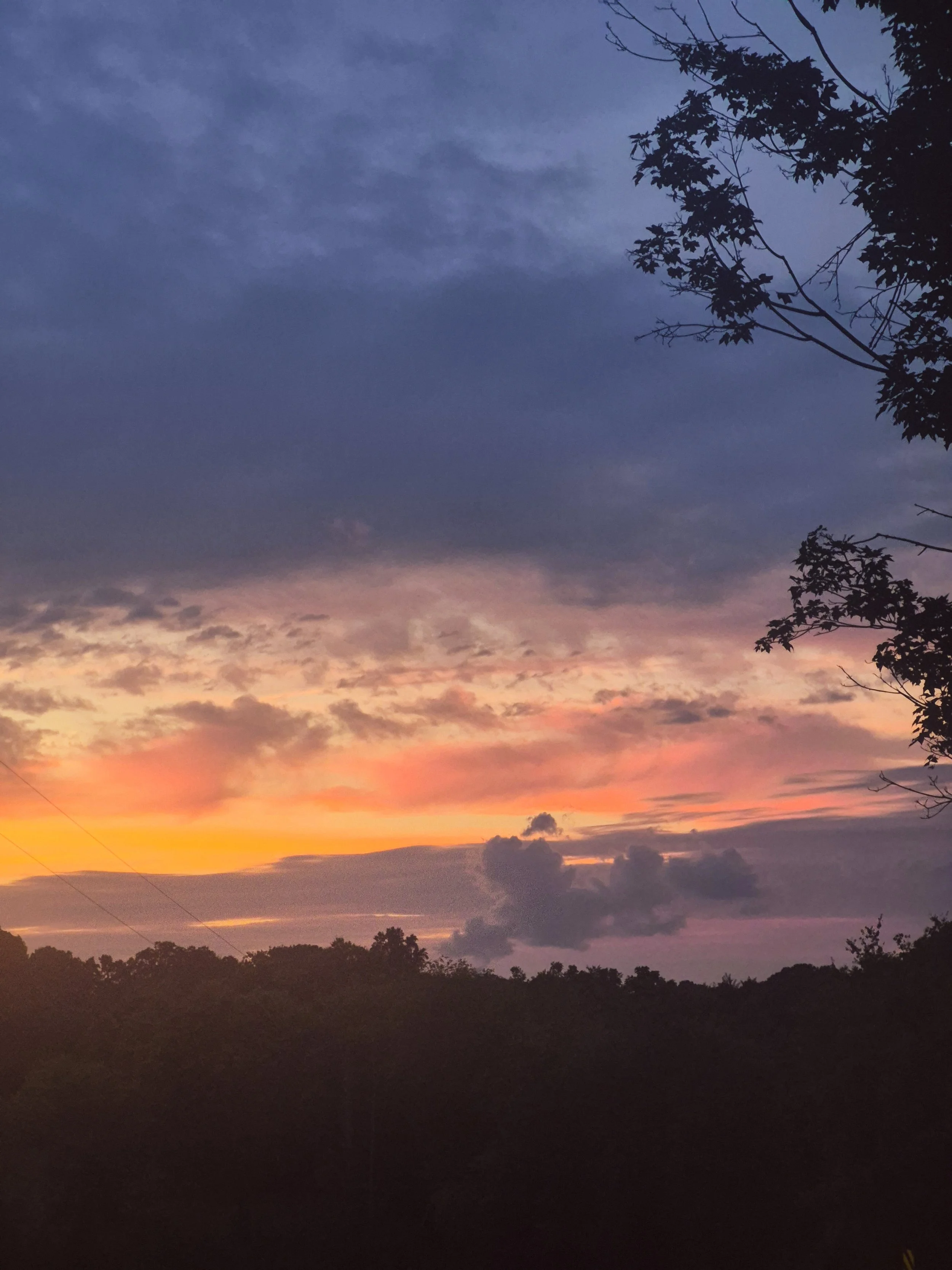 Dramatic sunset over a tree-covered landscape, with glowing orange and pink clouds beneath a deep blue evening sky.
