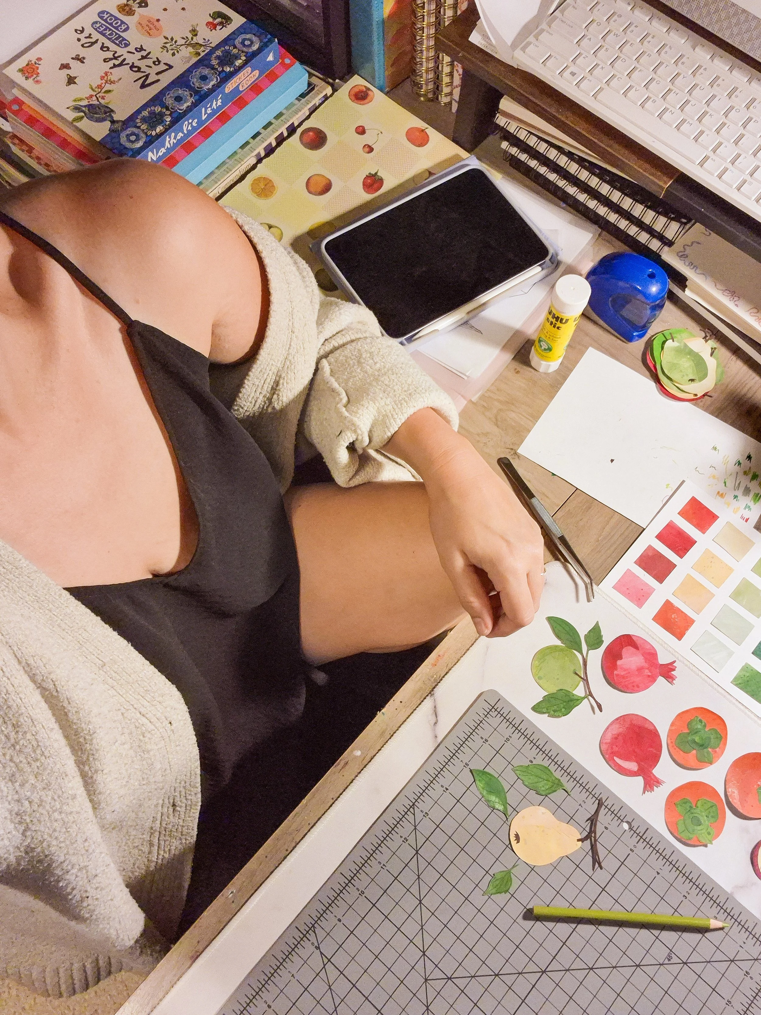 Artist working on hand-cut paper collage pieces at a studio desk surrounded by painted papers and art supplies..