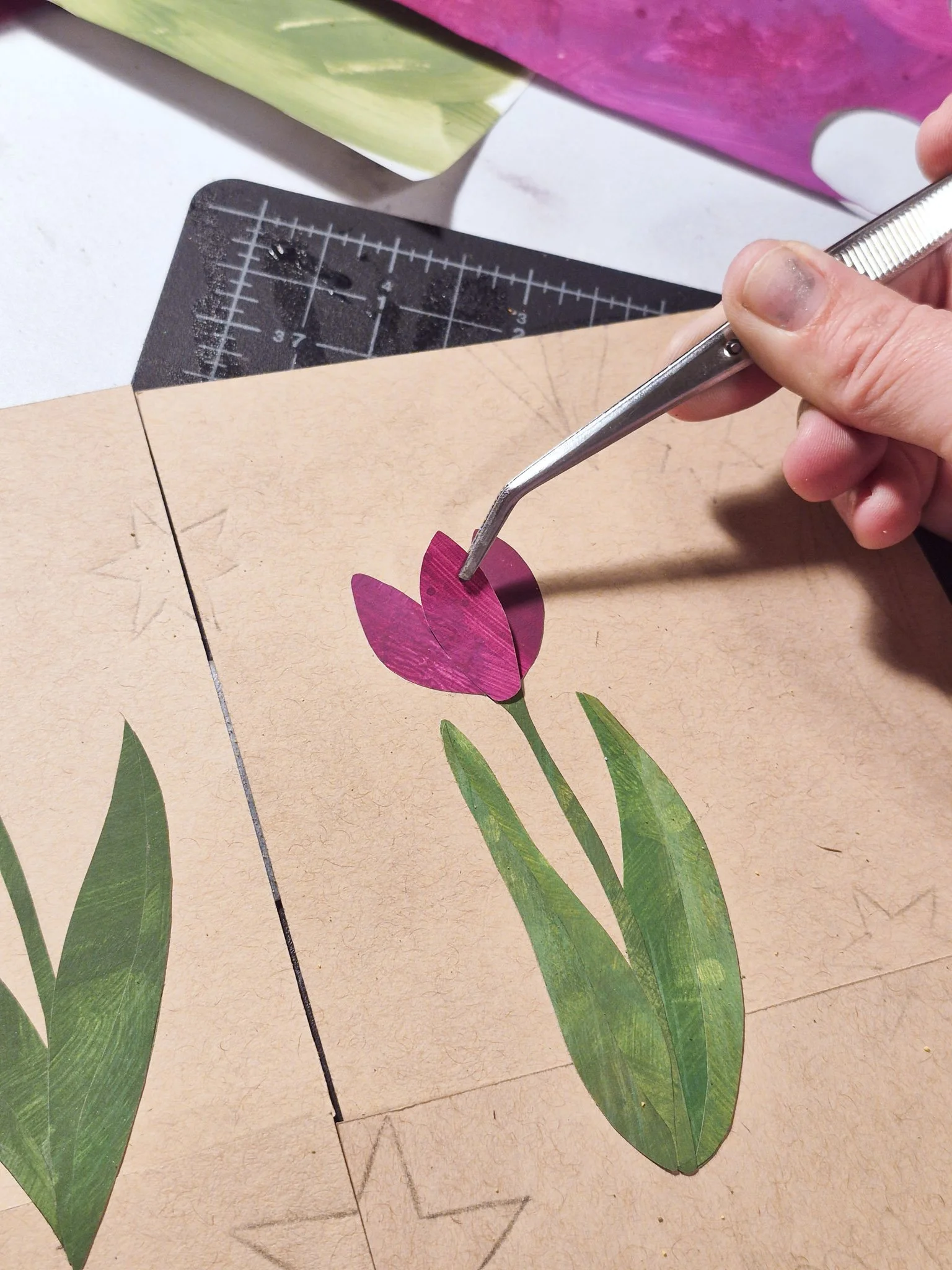 close up shot of a WIP painted paper collage tulip on a tan paper background. artist is adding a pink petal with tweezers