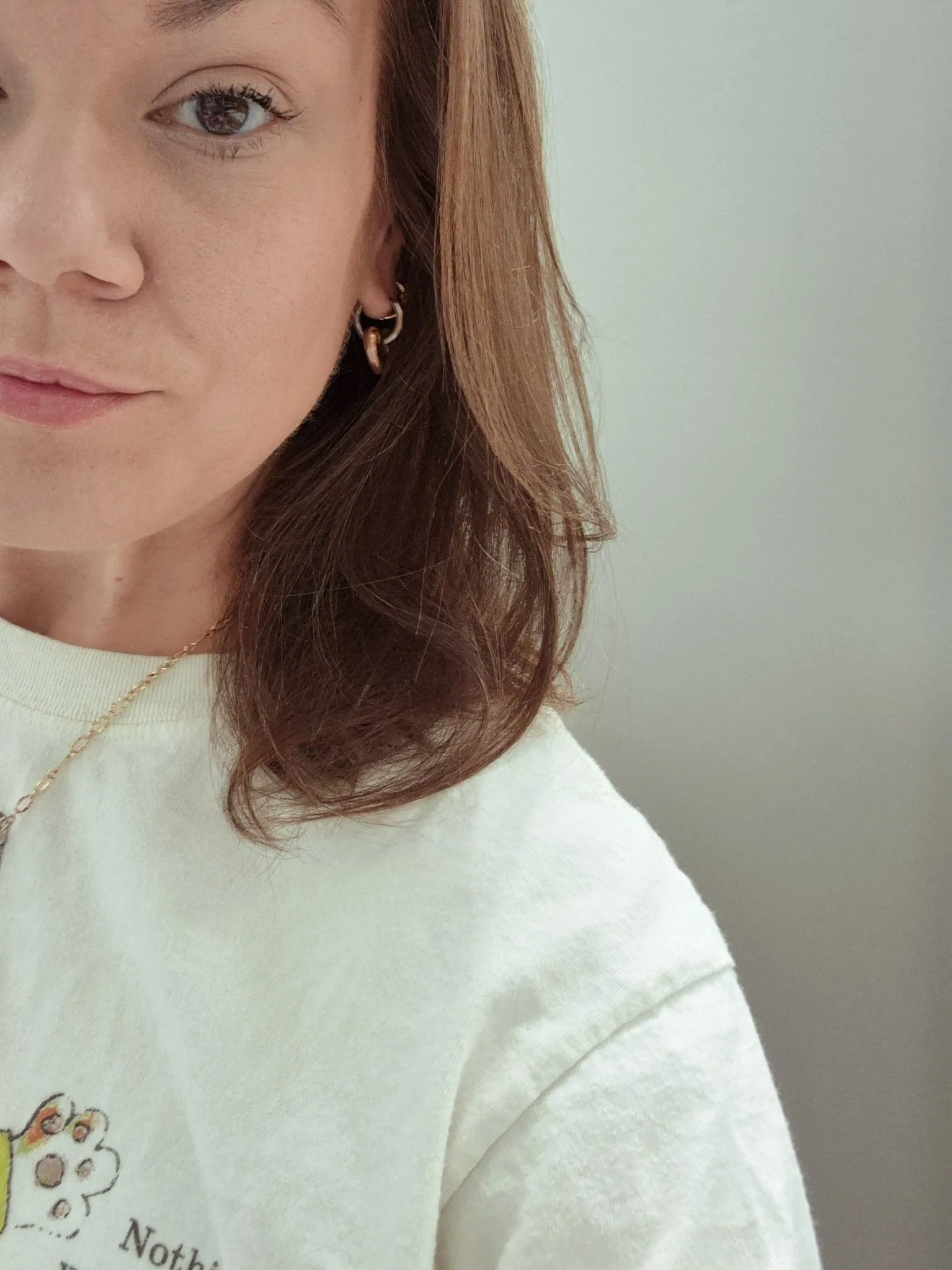 a selfie of the artist with only half her face in the frame of the image. shes wearing a neutral t shirt, gold earring and a gold necklace. she had mid-length brown bair and dark brown eyes