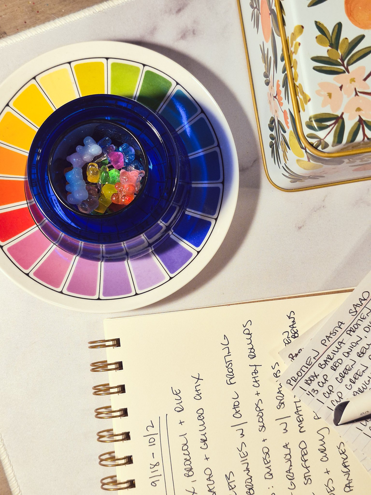 A color wheel trinket tray, colorful glass pieces, and a handwritten notebook resting on a studio worktable.