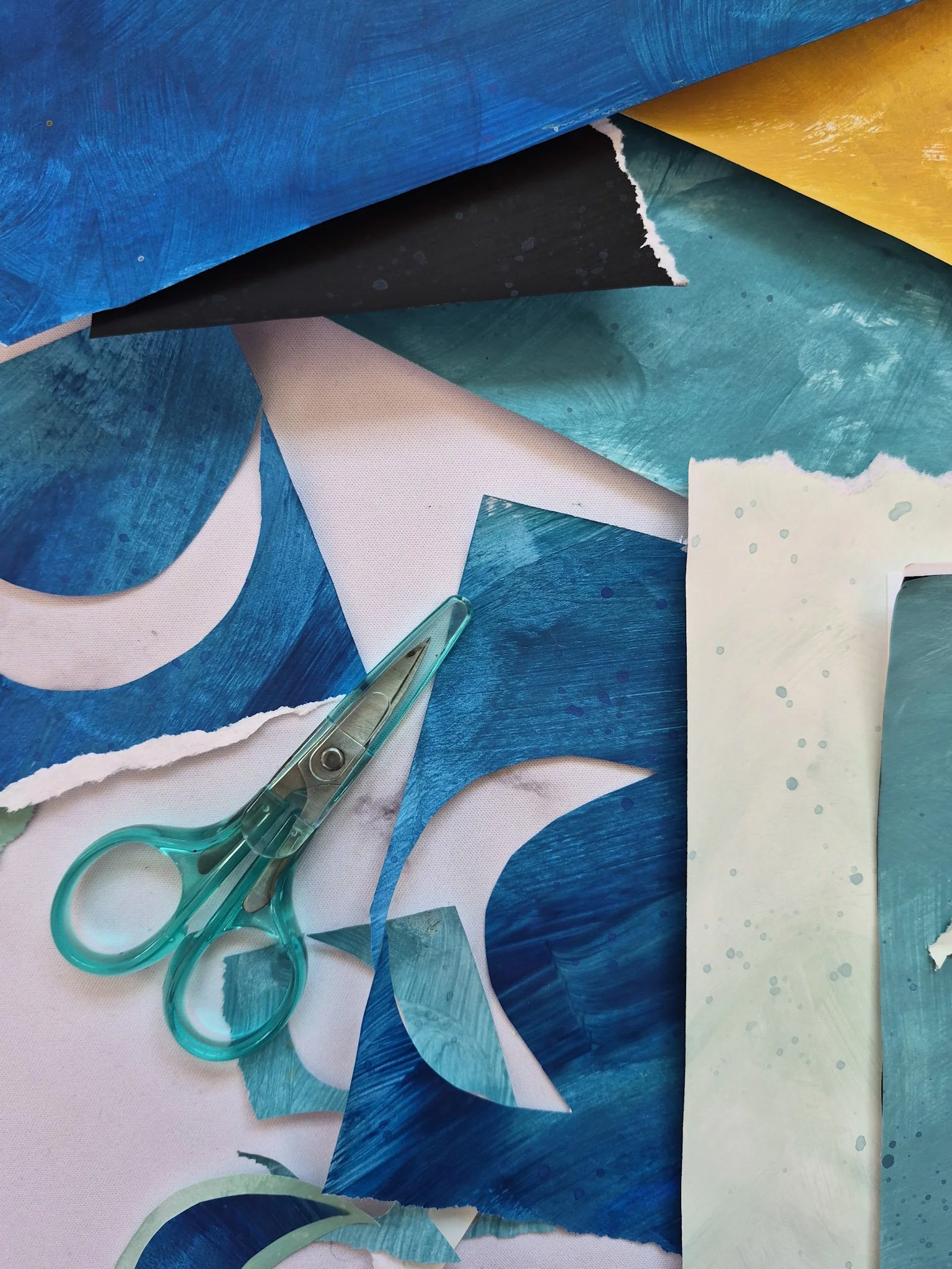 overhead shot of desk with painted papers of blues, aquas, and golds with scissors nearby