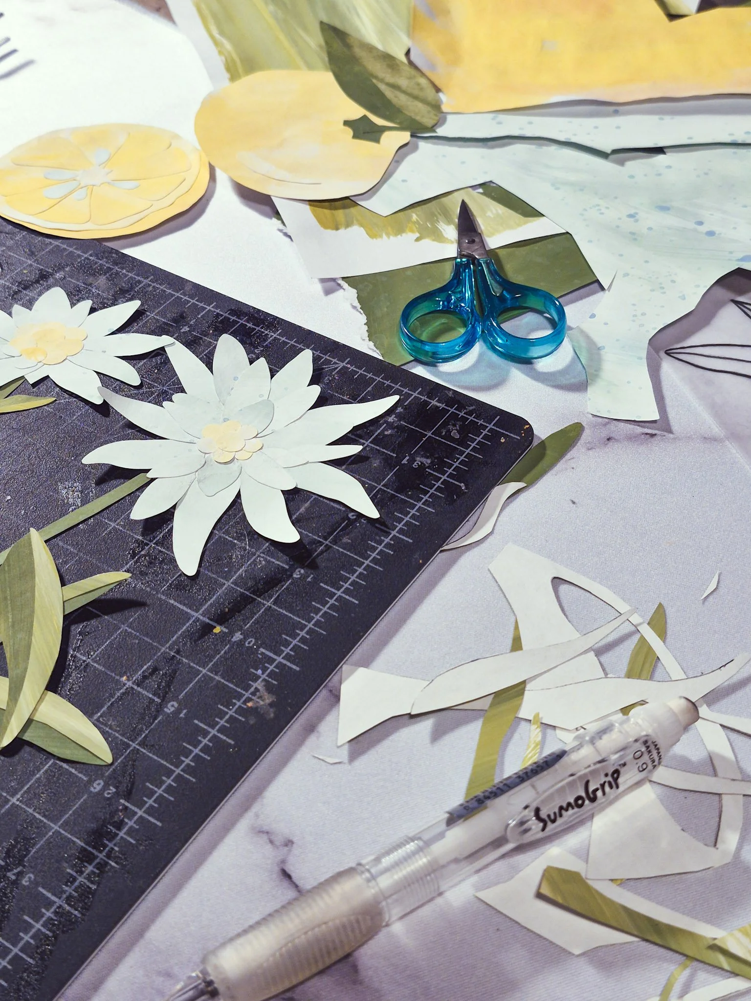 process photo of painted paper collage illustration with bits of paper a cutting mat scissors and a Sumo pencil on a white marble background