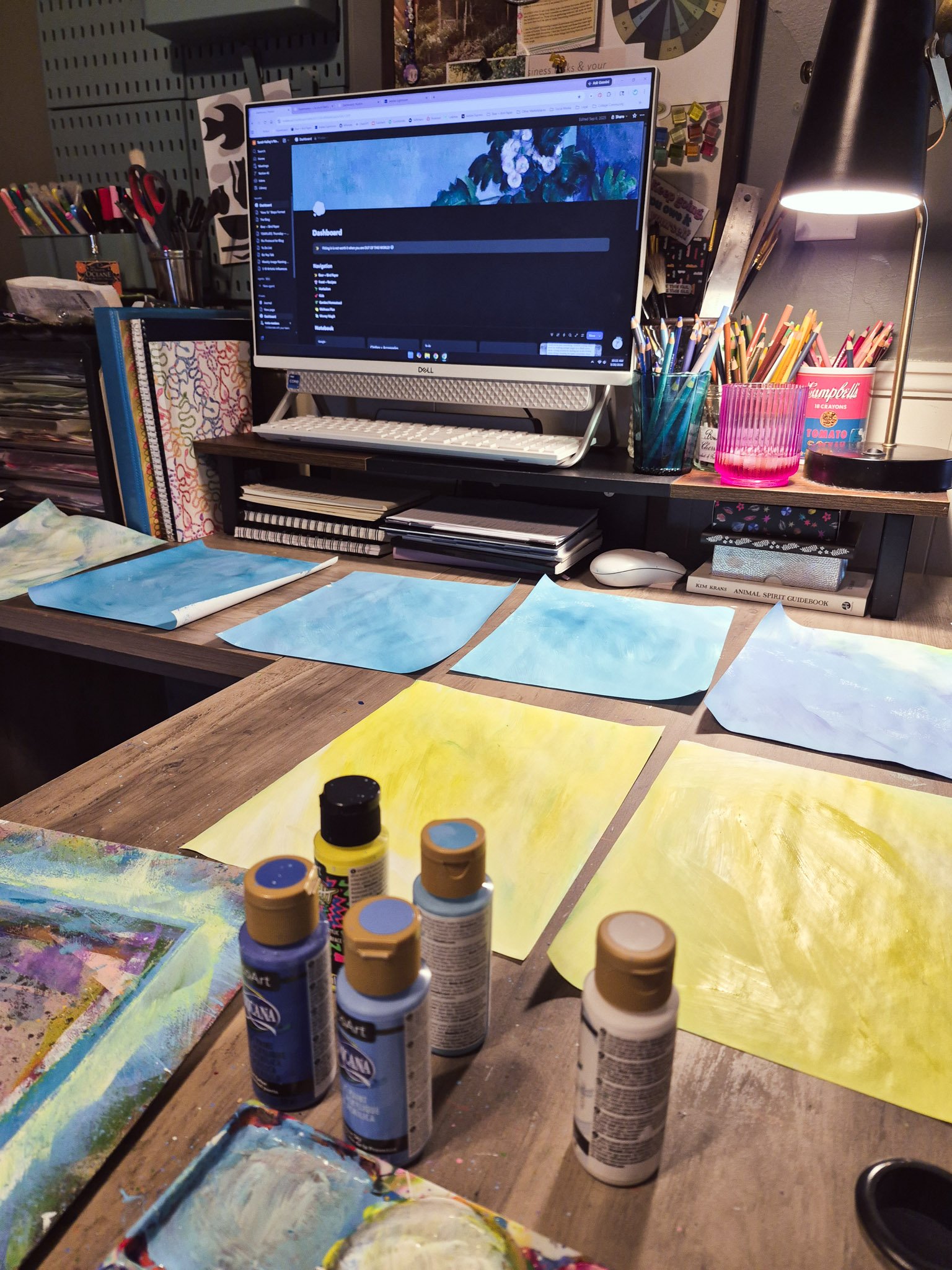 Cozy art workspace with a desk covered in hand-painted blue and yellow paper backgrounds, acrylic paint bottles in the foreground, and a computer and drawing supplies in the background.