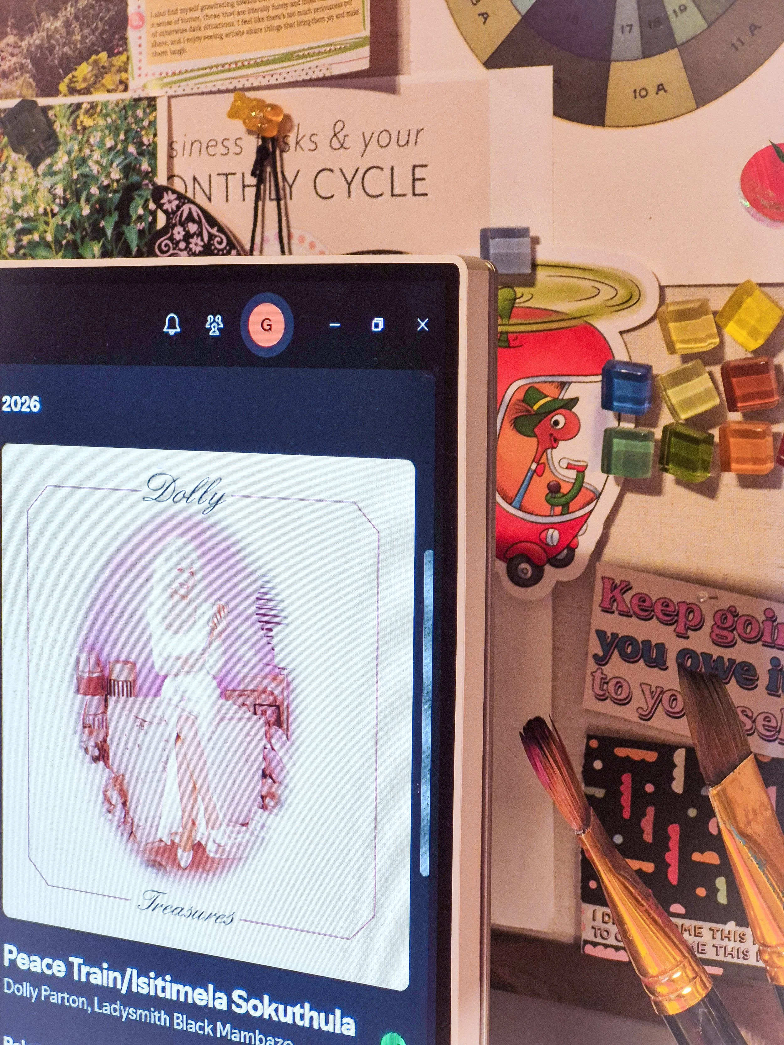 Studio desk with an iPad displaying Dolly Parton artwork, surrounded by paintbrushes, colorful paper collage pieces, and handwritten creative notes.