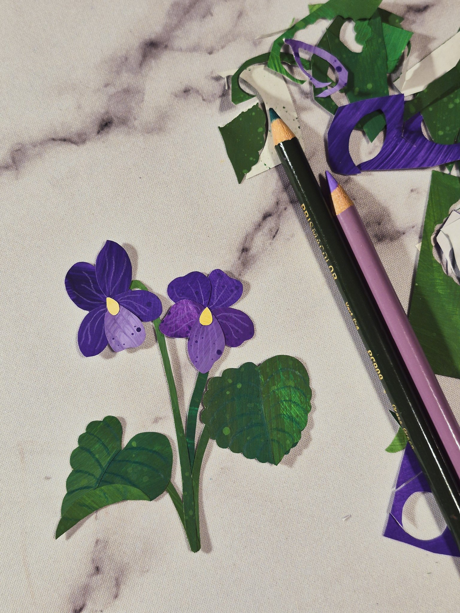 a close up desk shot of a painted paper collage violet and leaves on a marble background with art supplies around the desk