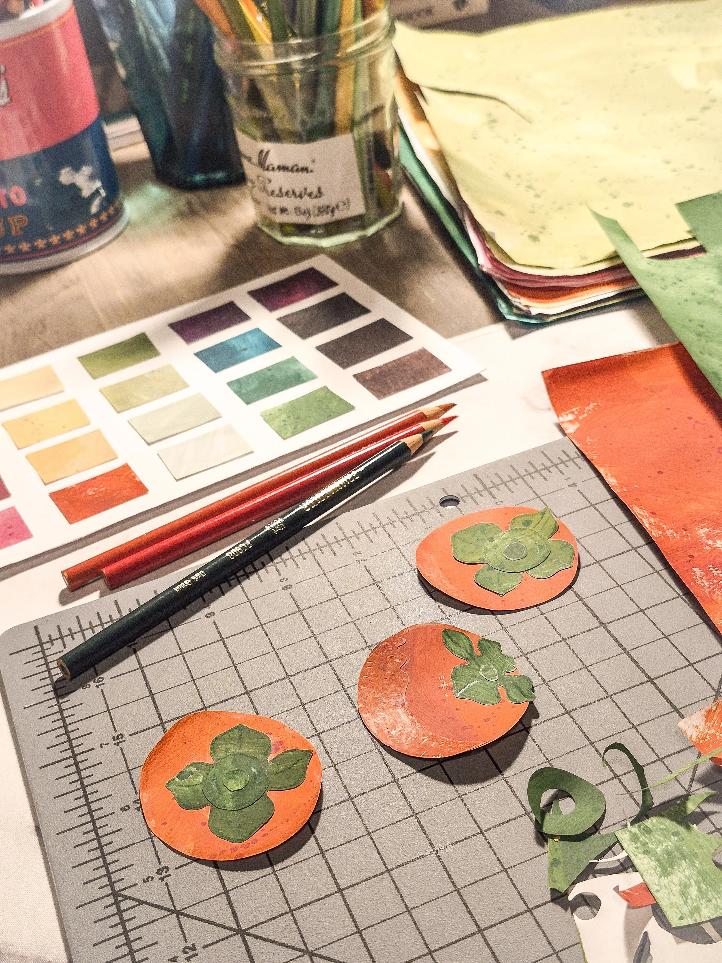 Painted paper persimmons arranged on a cutting mat with colorful painted paper in the background