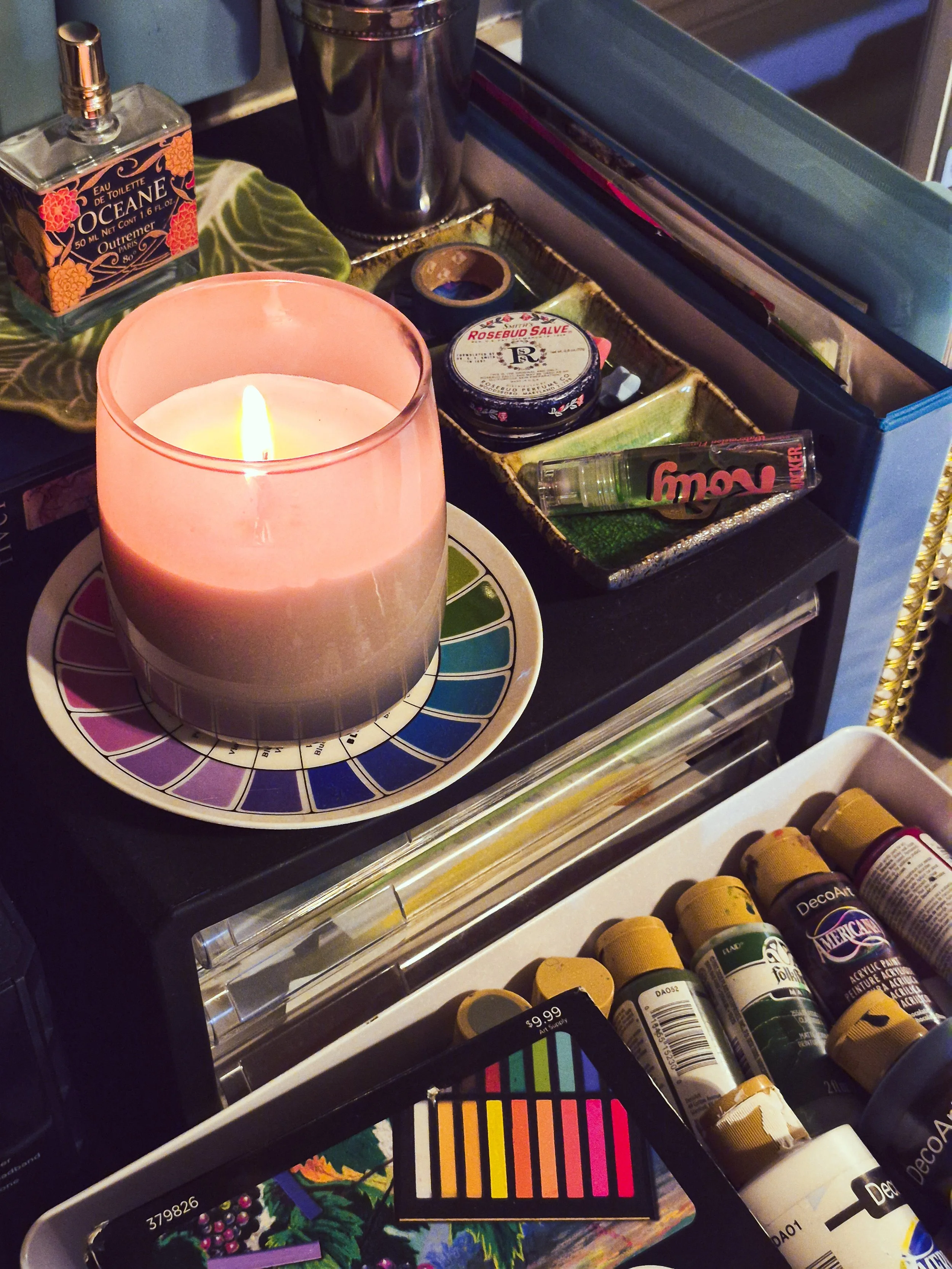 Studio storage drawer filled with acrylic paints and art supplies beside a lit candle, color swatches, and vintage objects.