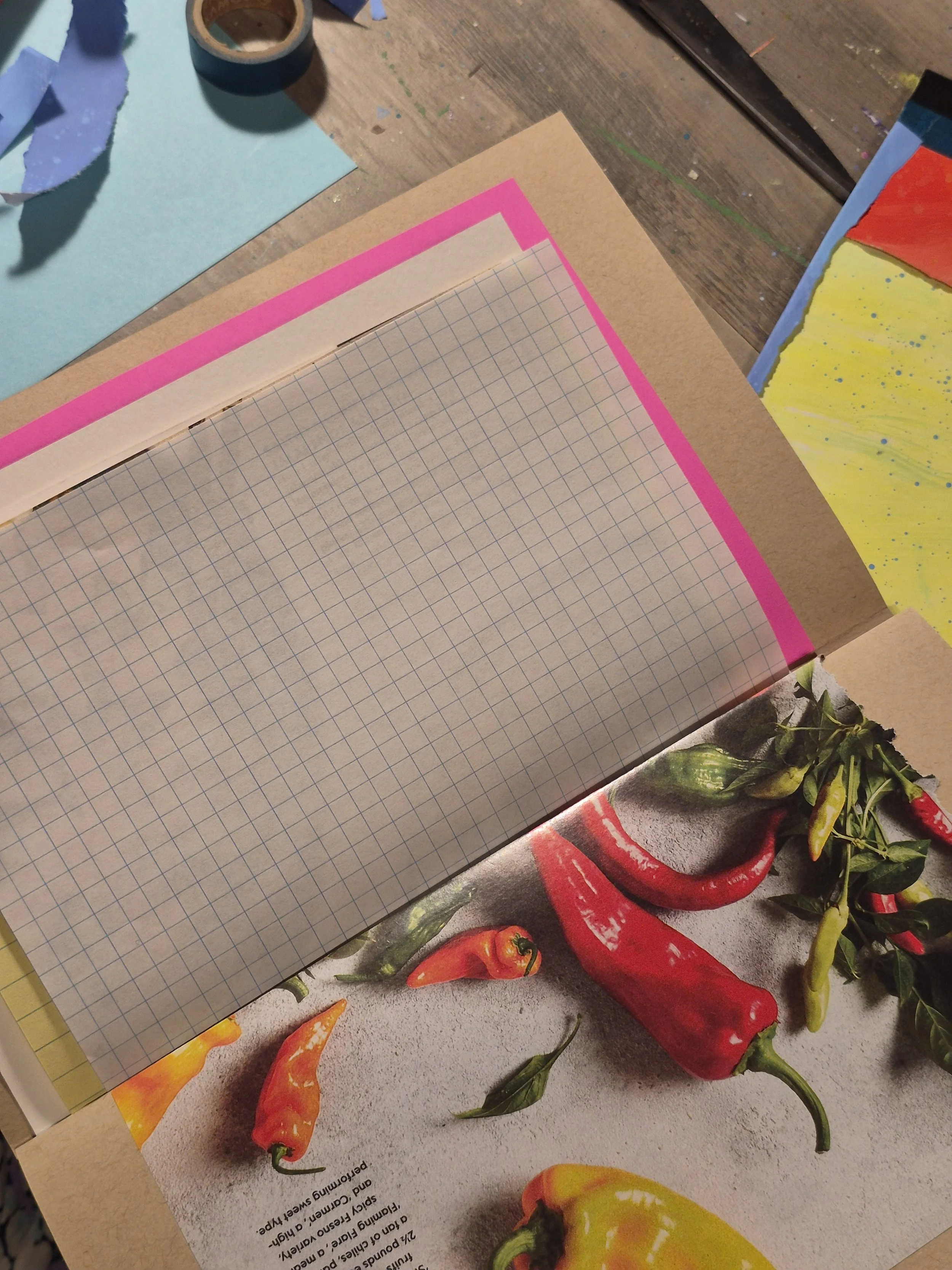 Artist’s worktable with a graph-paper sketchbook, painted papers, masking tape, and colorful art materials beside a vintage cookbook page featuring peppers.