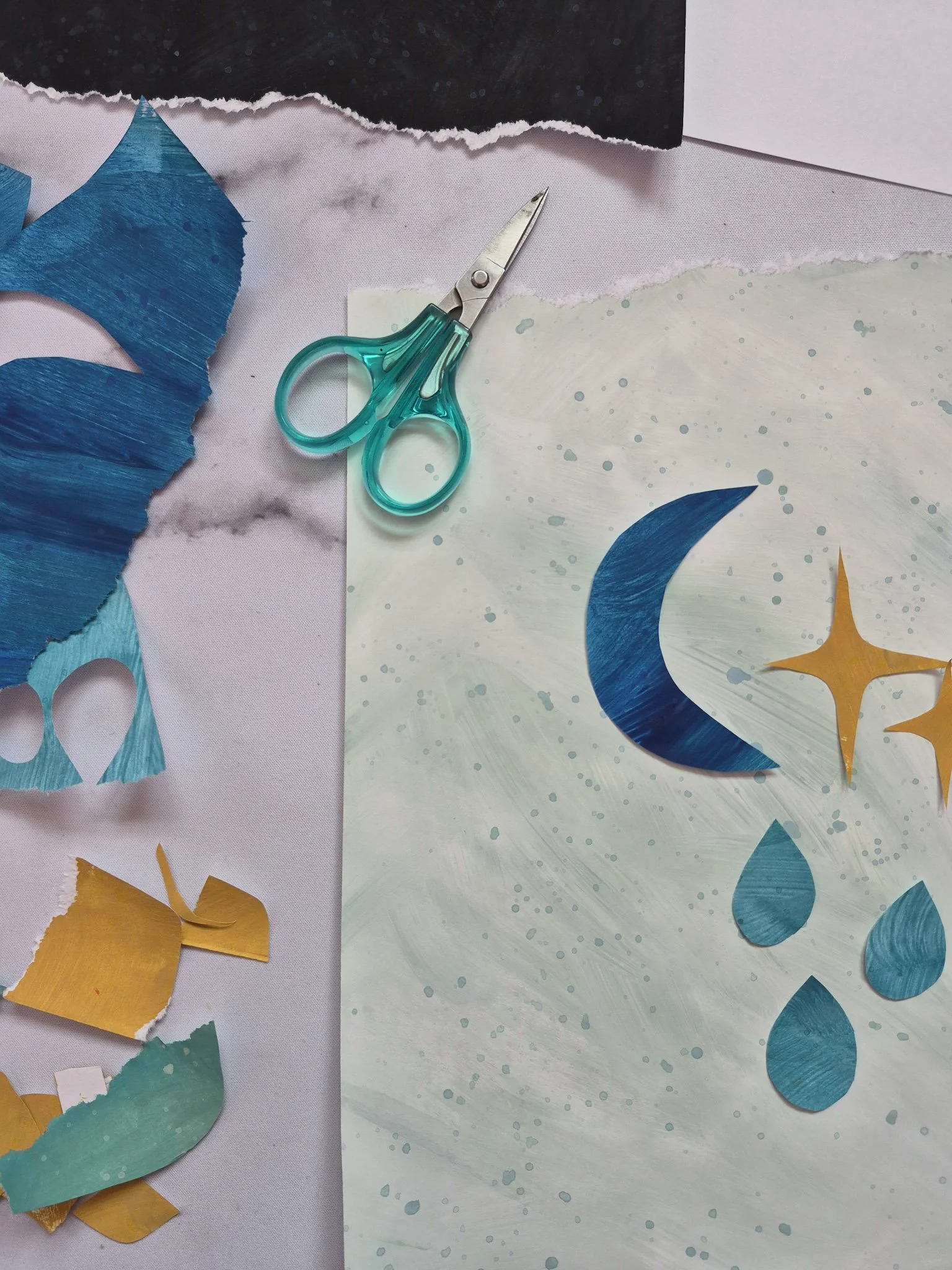 overhead desk shot of hand painted papers being cut for collage. crescent moon, raindrops and retro starbursts