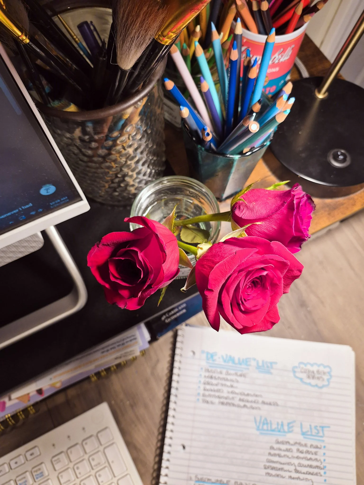 cozy desk shot with a jam jar holding three red roses also various pots containing colored pencils and art supplies