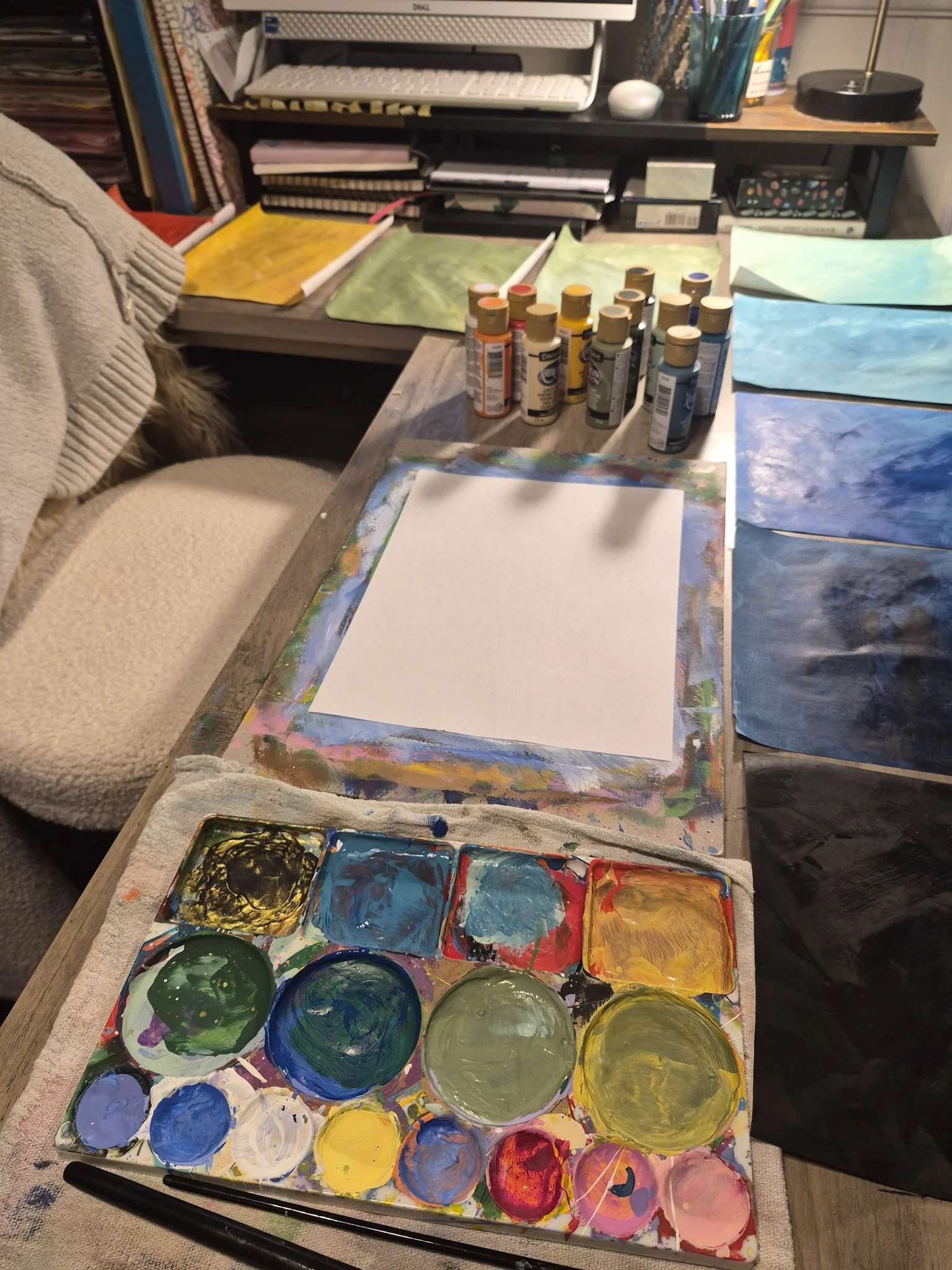 workspace shot of a desk with paints, a palette, and painted papers all around