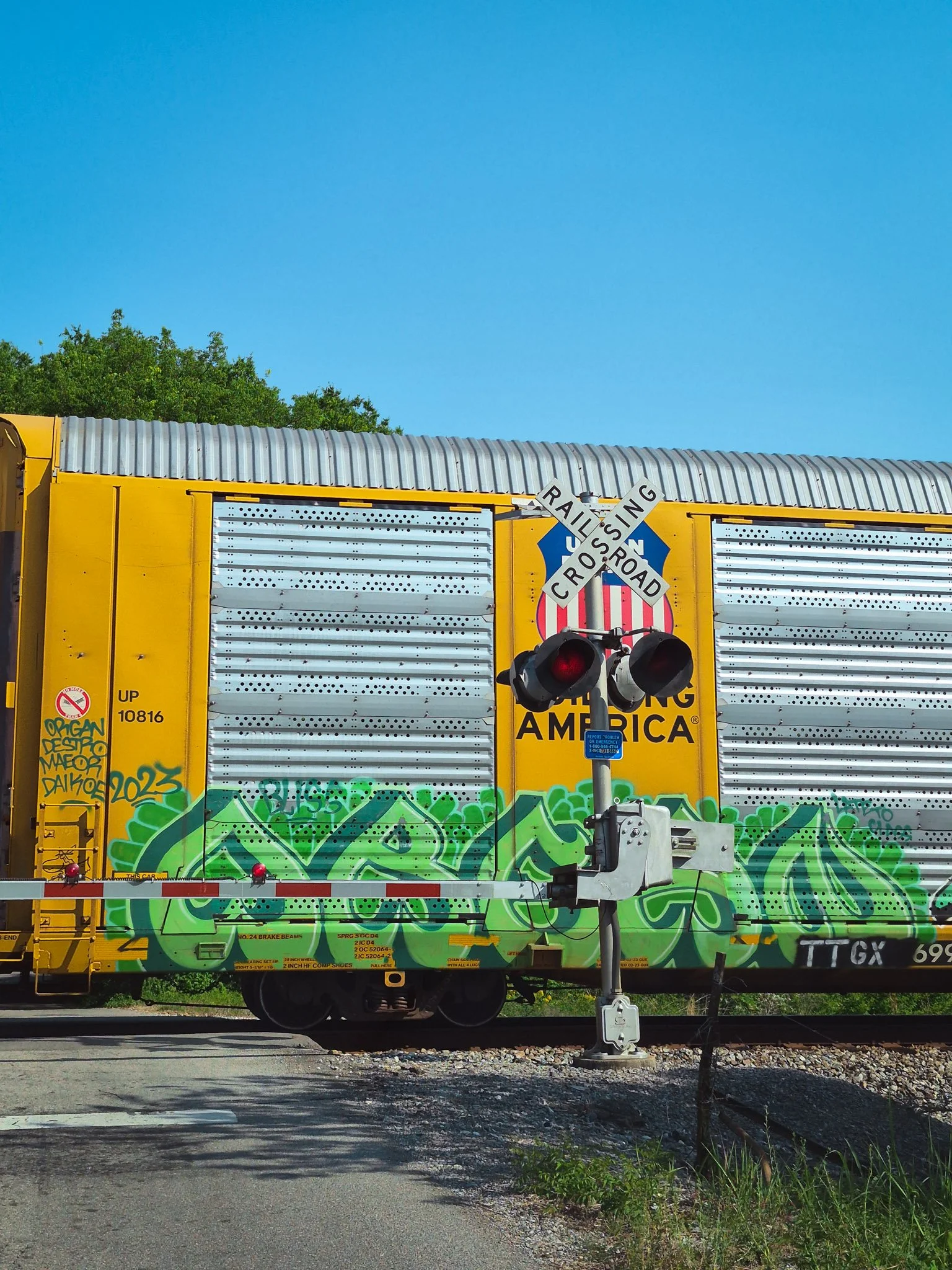 Yellow freight train car with green graffiti moving past a railroad crossing sign.