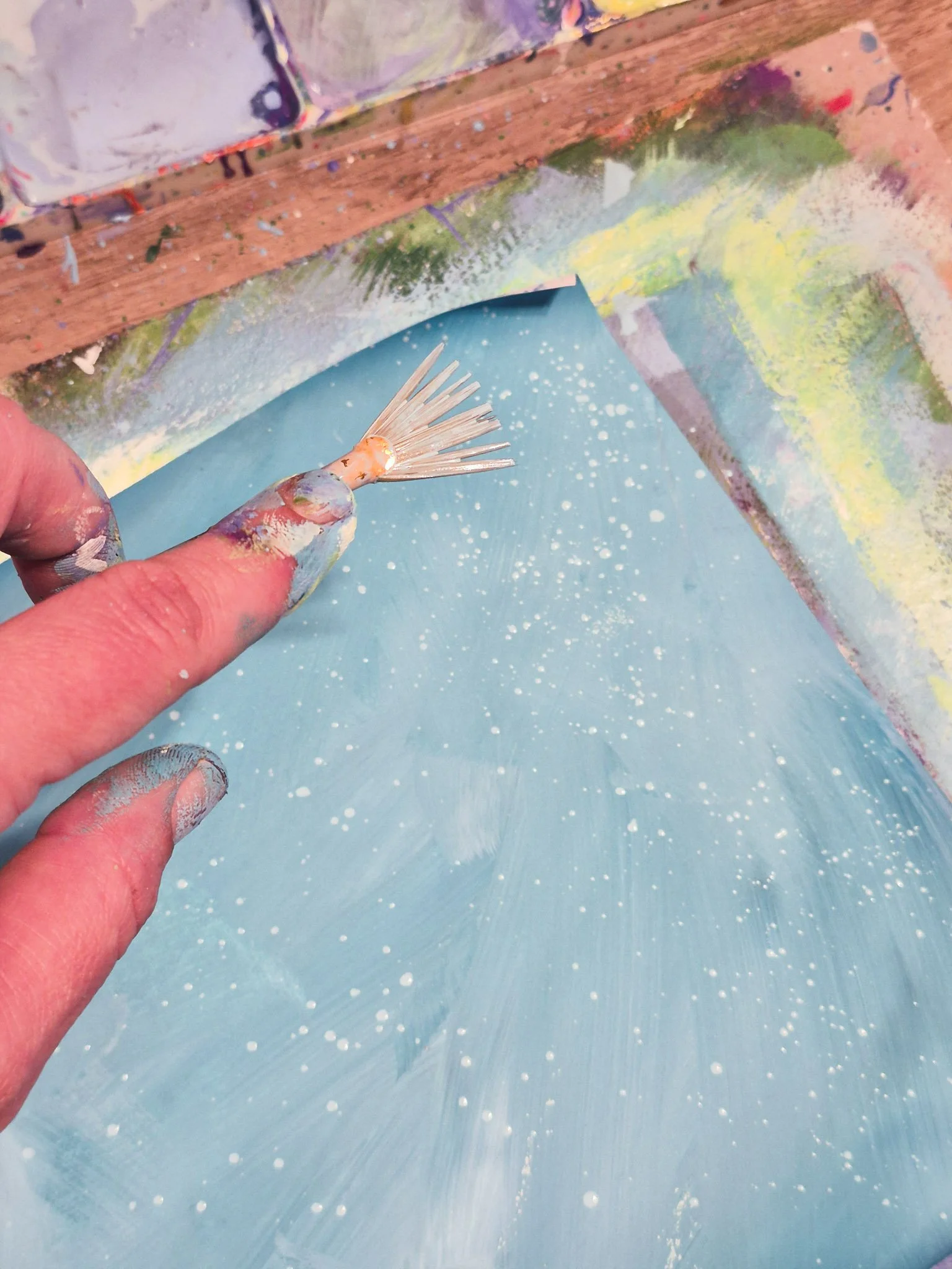 closeup view of a pale blue, hand-painted paper with the artist's hand holding a paintbrush above it, splattering a fine spray of a lighter blue paint, adding texture.