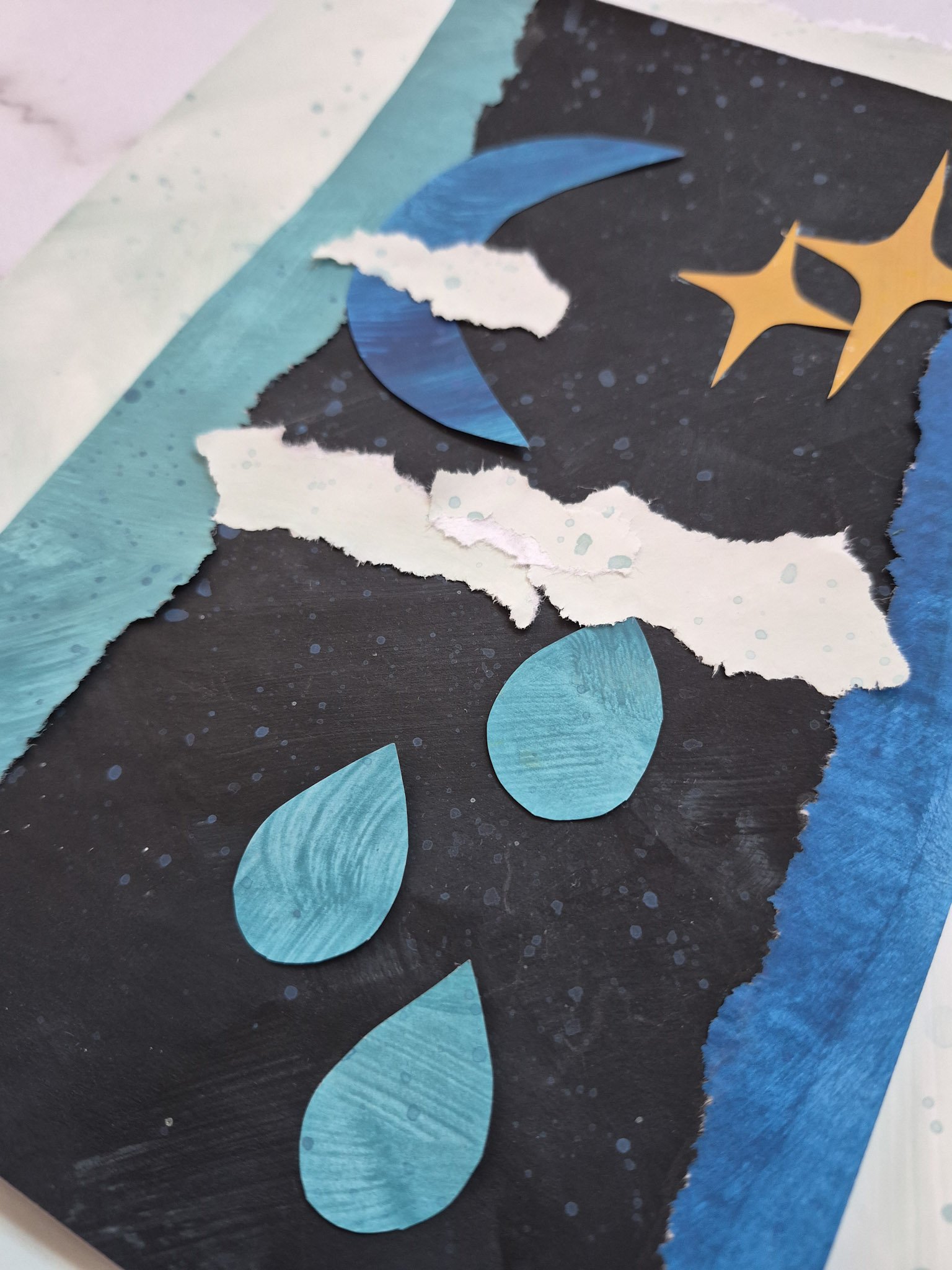 shot of final collage with crescent moon, clouds, raindrops and starbursts on a black painted paper background that resembles outerspace