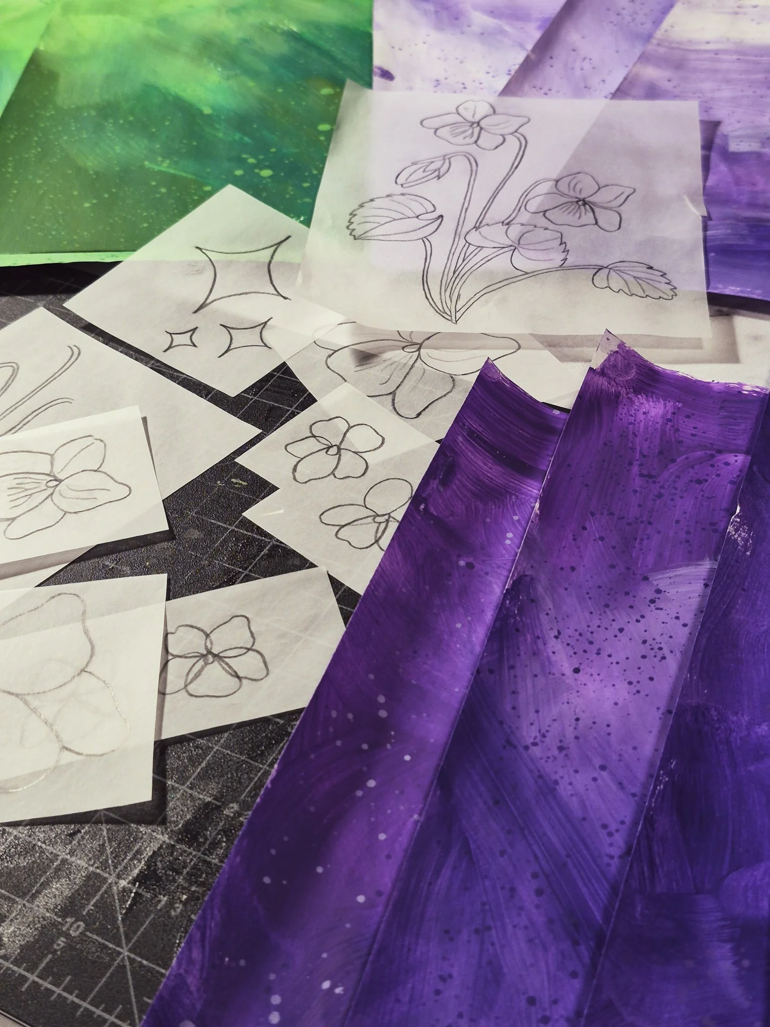 an overhead view of a desk covered in painted collage papers in shades of emerald green and violet with a translucent tracing paper template of a wild violet plant sitting on top.
