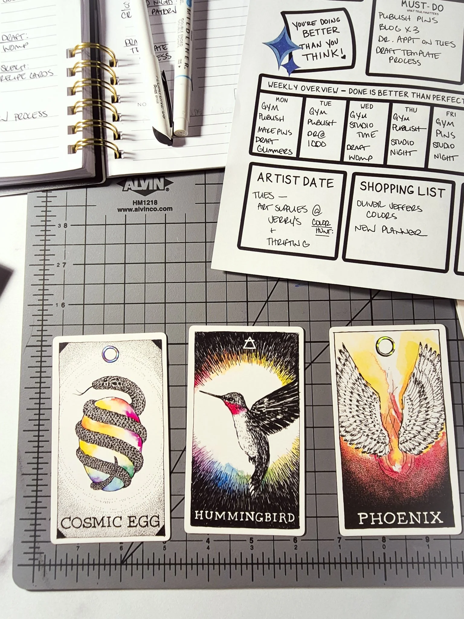 Illustrated oracle cards (cosmic egg, hummingbird, phoenix) styled with a weekly creative reset planner, pens, and notebook on a grid cutting mat desk setup