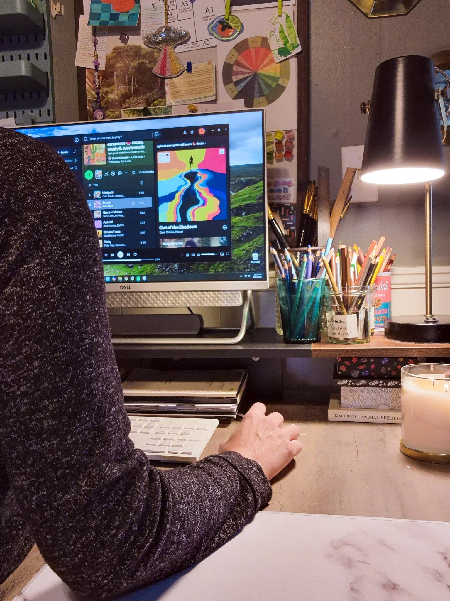 an image of the artist in her studio,  sitting at her computer working on illustrations. workspace is decorated with jars and jars of art supplies, brushes and markers and a large computer screen as well as vision boards filled with art and inspo
