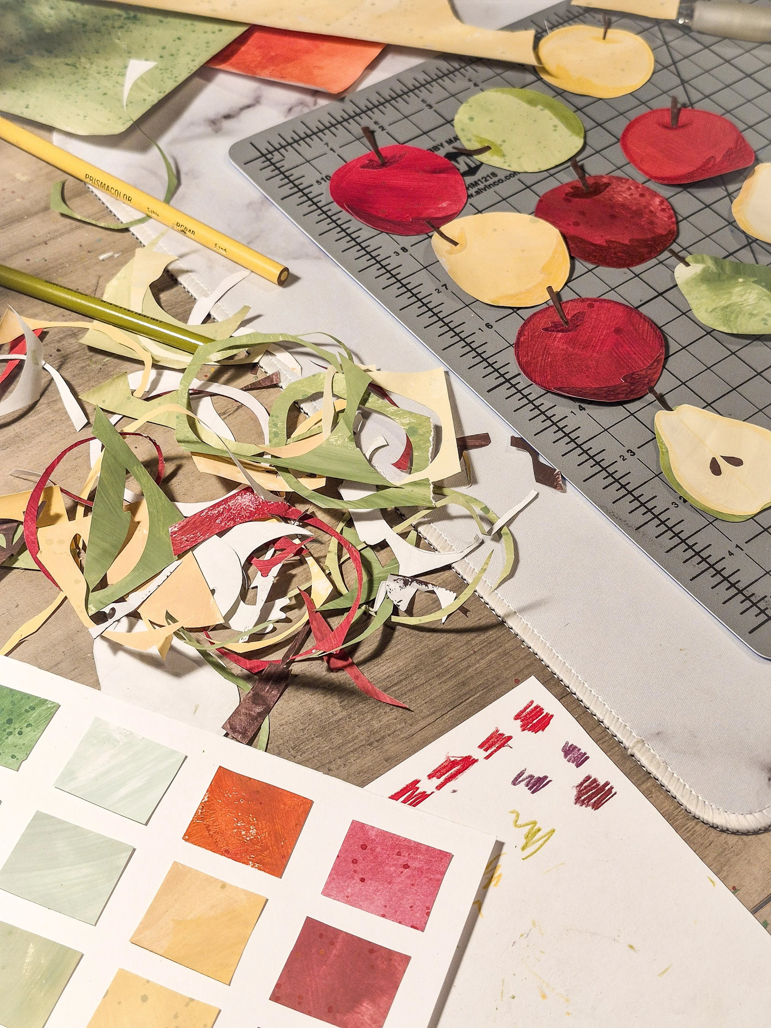 Hand-painted paper apple shapes arranged on a cutting mat beside colored pencils, paint swatches, and collage scraps.