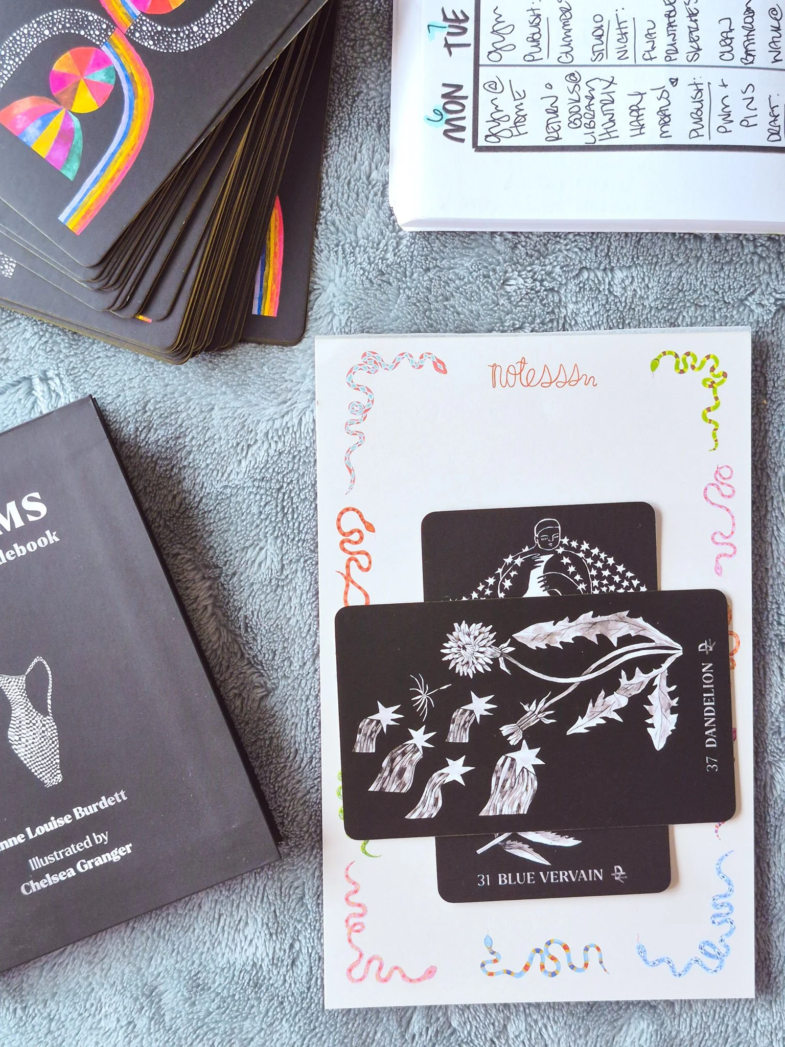 A cozy oracle card and planning setup with botanical oracle cards (dandelion and blue vervain), a weekly planner spread, and illustrated ‘Dirt Gems’ deck styled on a soft blanket with natural, earthy tones.