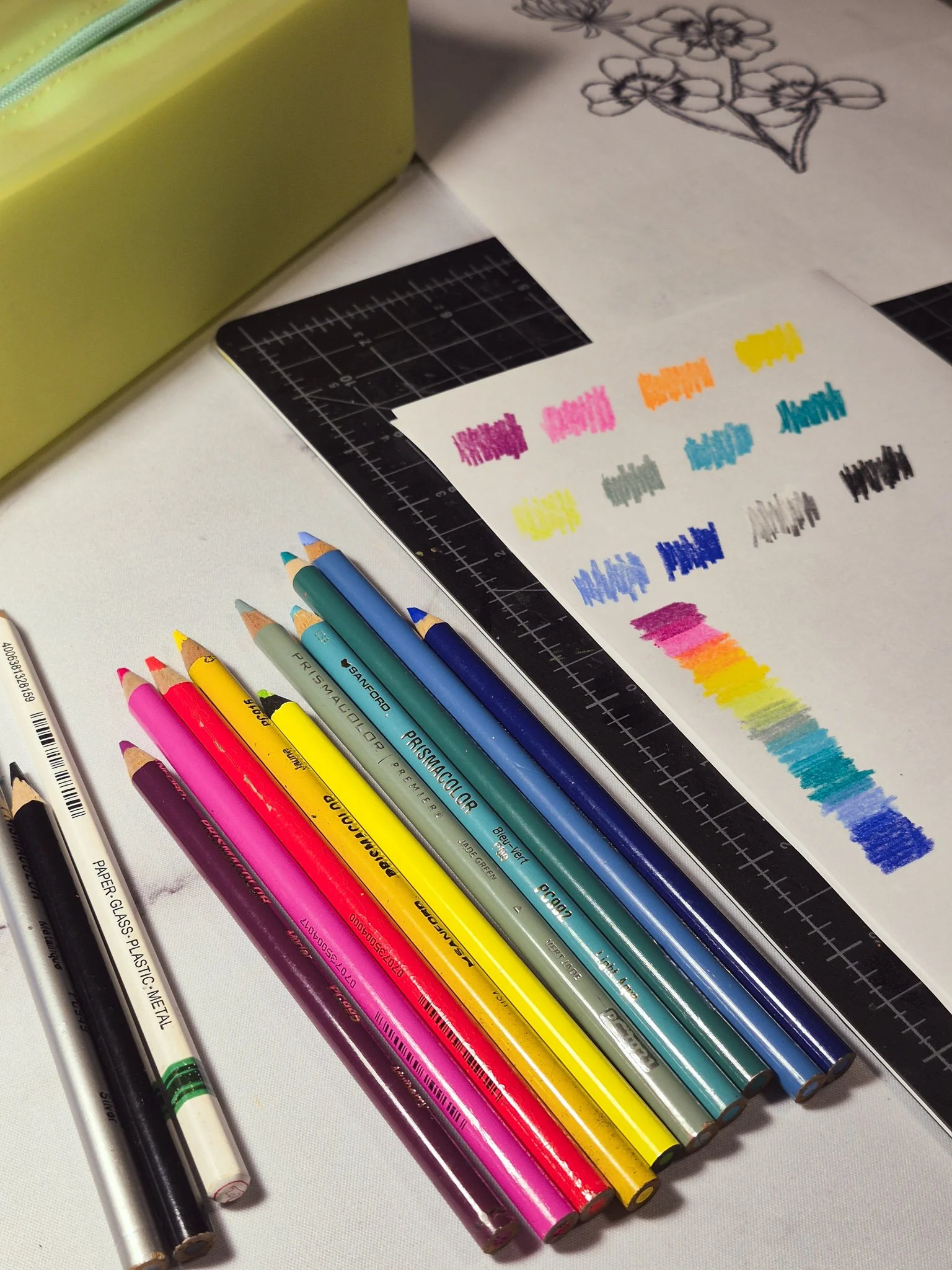 a curated set of Prismacolor colored pencils with pops of neon set next to a black cutting mat and a pencil sharpener along with a piece of paper scribbles with colors