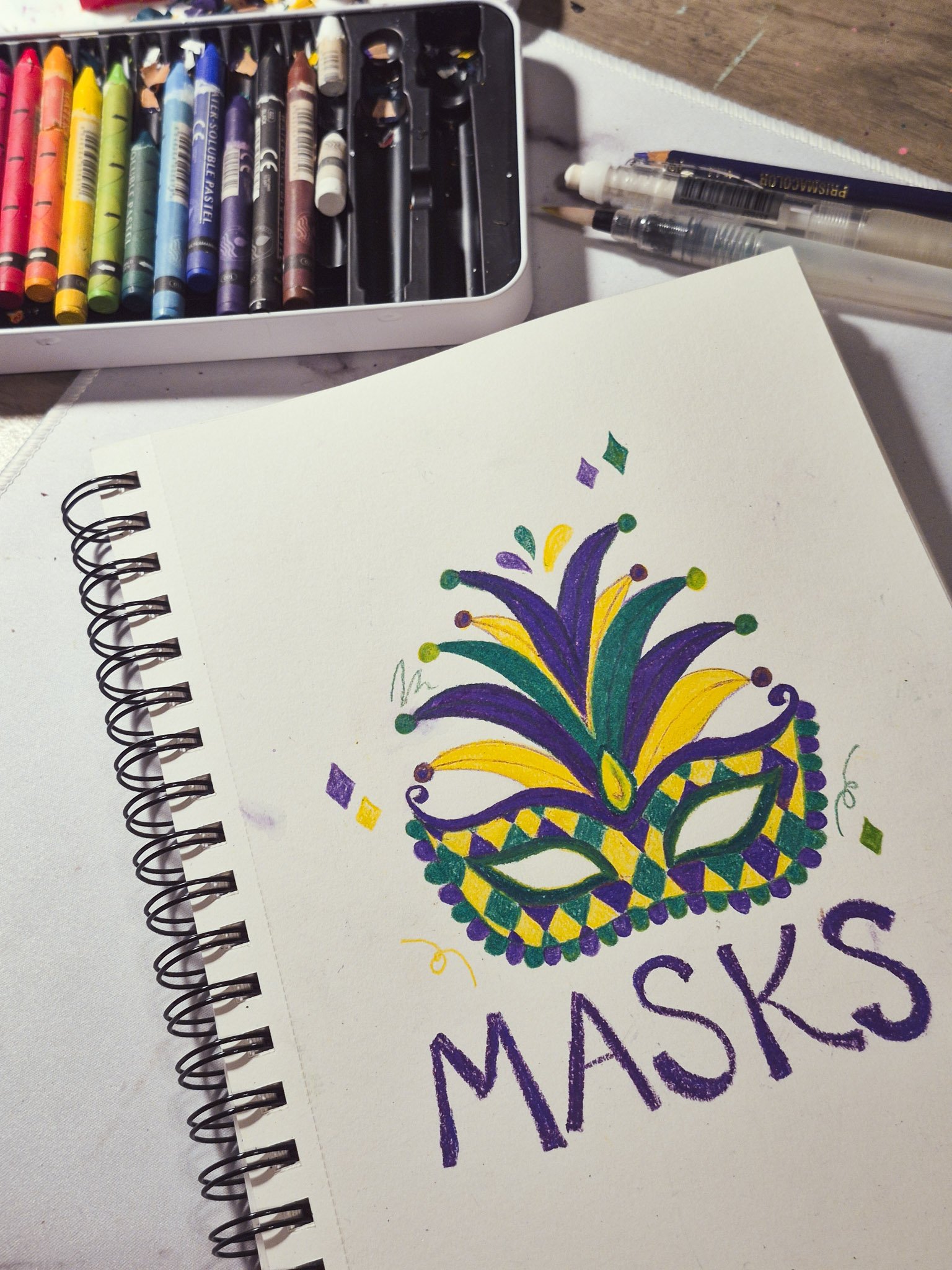 overhead desk shot of a sketchbook with an illustration of a mardi gras mask and