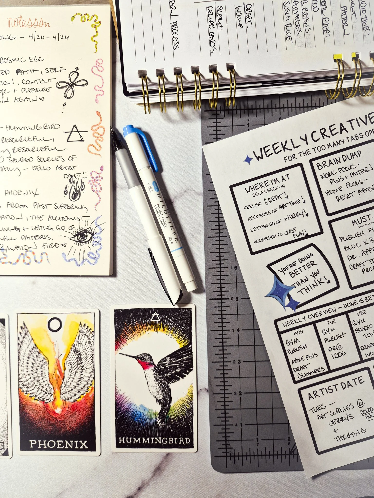 Weekly creative reset planner sheet with handwritten notes, pens, sketchbook doodles, and phoenix and hummingbird oracle cards in a cozy creative workspace