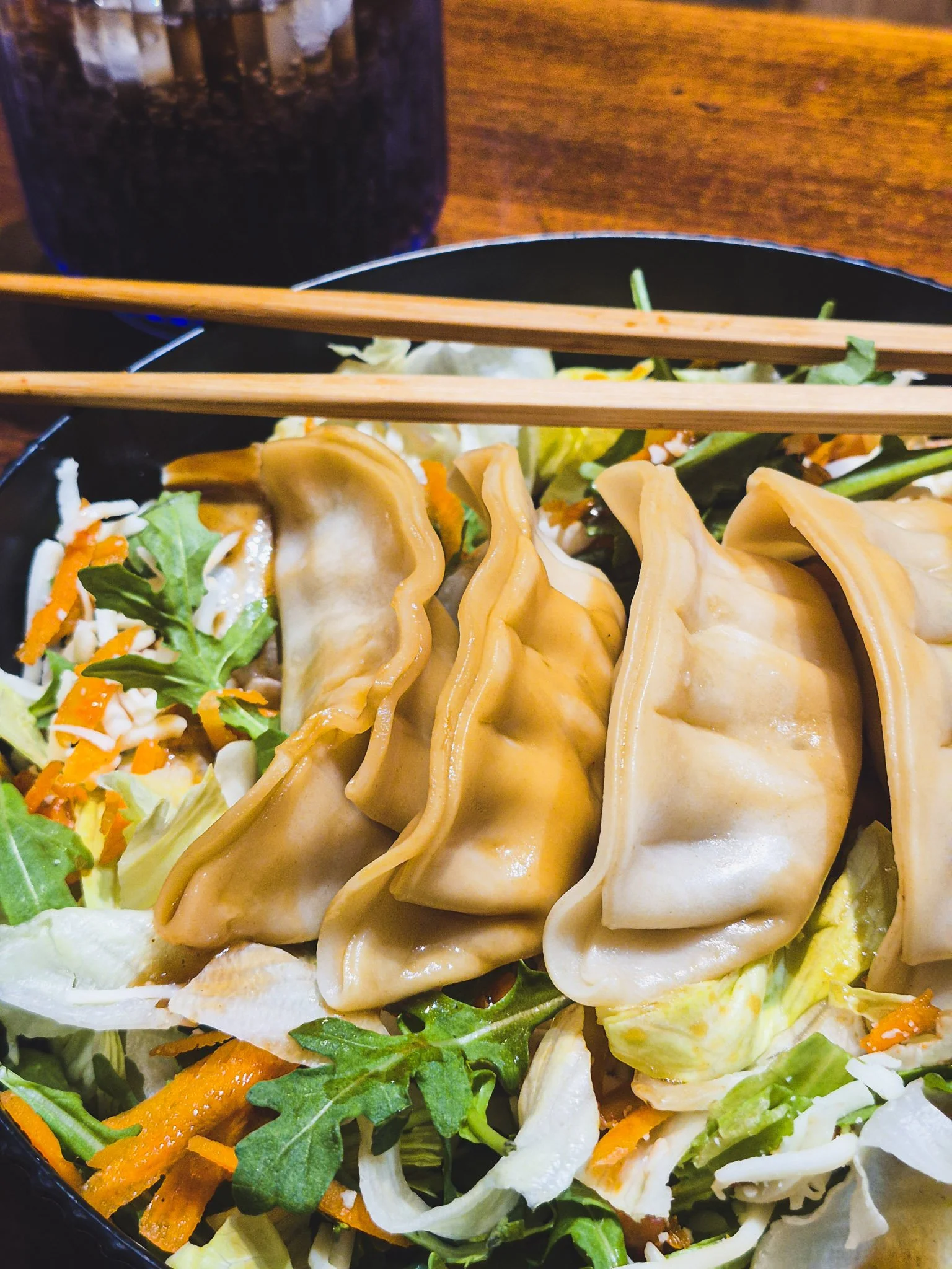 a closeup of a black bowl with chopsticks laid across it containing a dumpling salad with arugula, carrots, sesame dressing, iceberg, dumplings, and Fly By Jing chili crisp