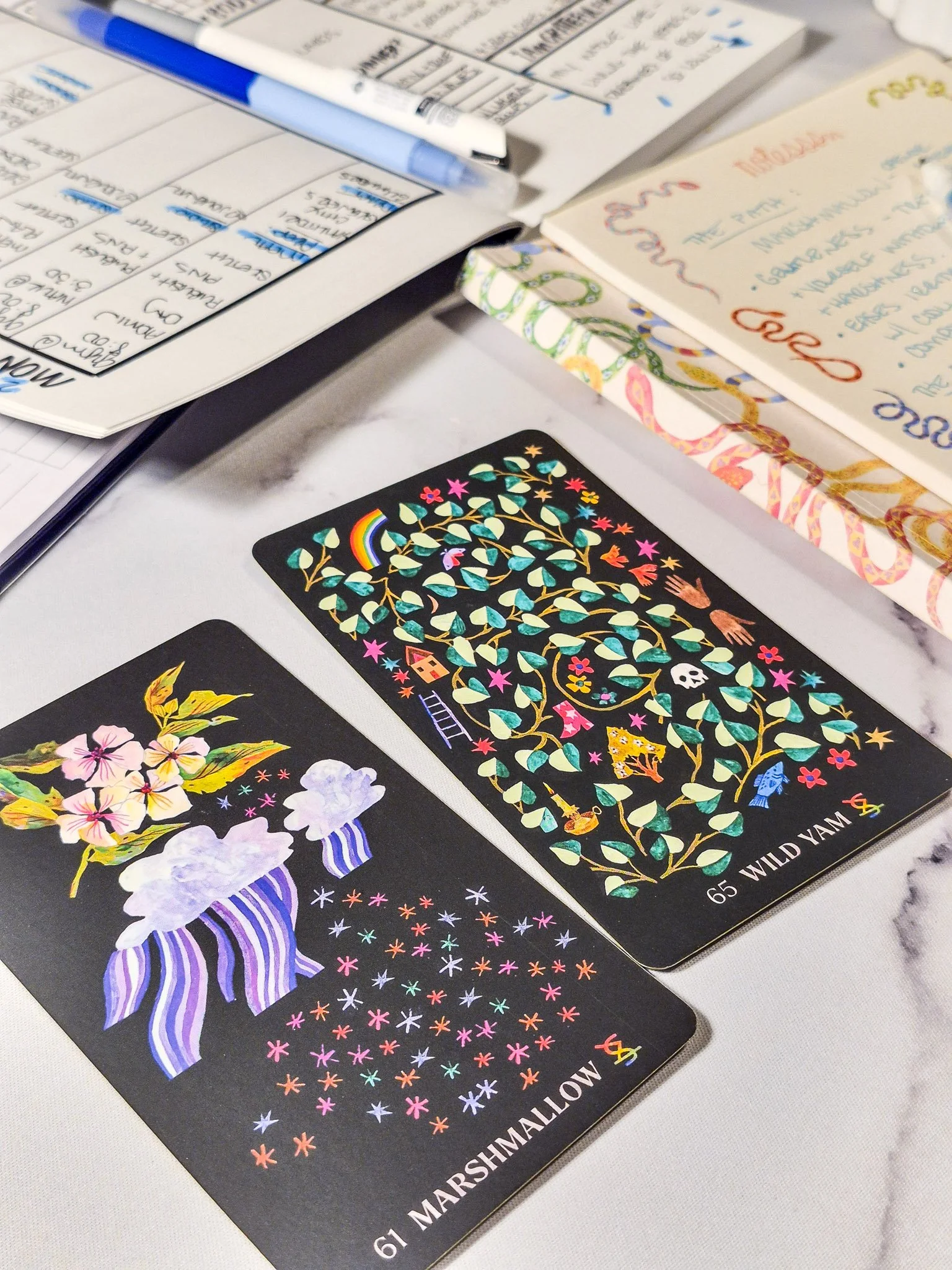 Illustrated tarot-style art cards beside a handwritten weekly planner spread and blue pens on a marble surface, cozy stationery setup