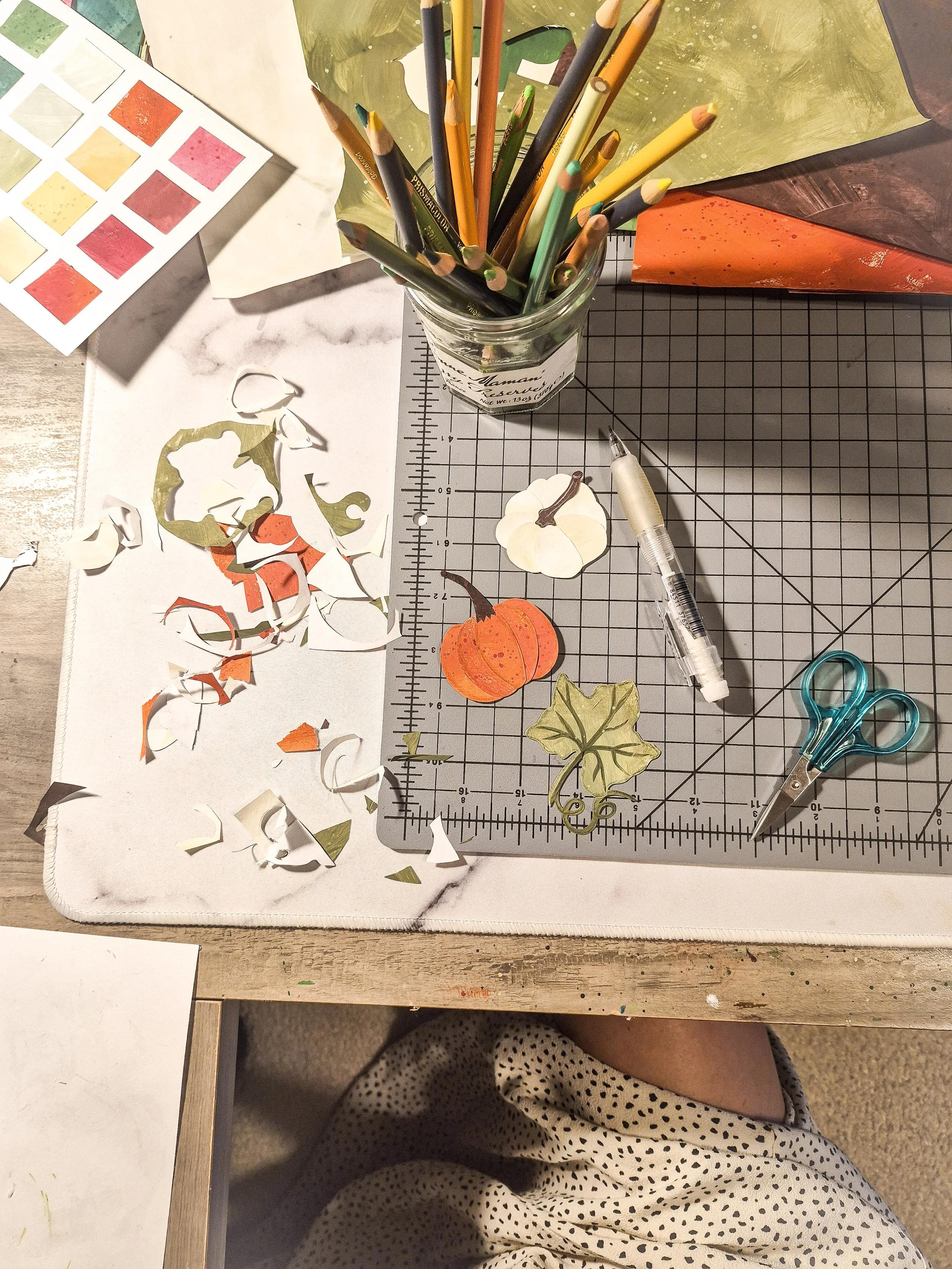 Hand-cut painted paper pumpkin pieces and leaves arranged on a cutting mat in the studio.