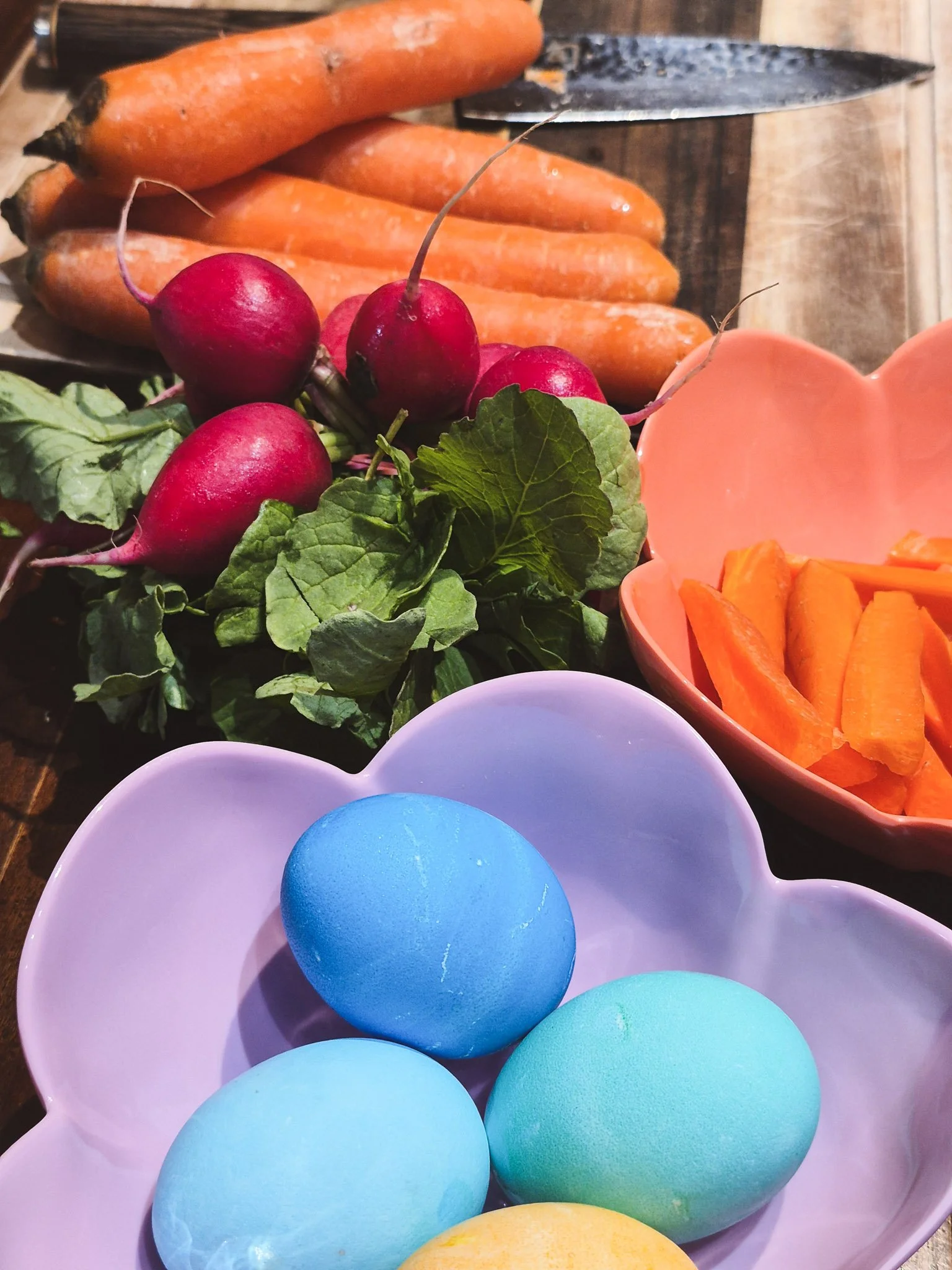 closeup image of fresh spring produce near a cutting board and pastel flower bowls containing crudités and dyed easter eggs