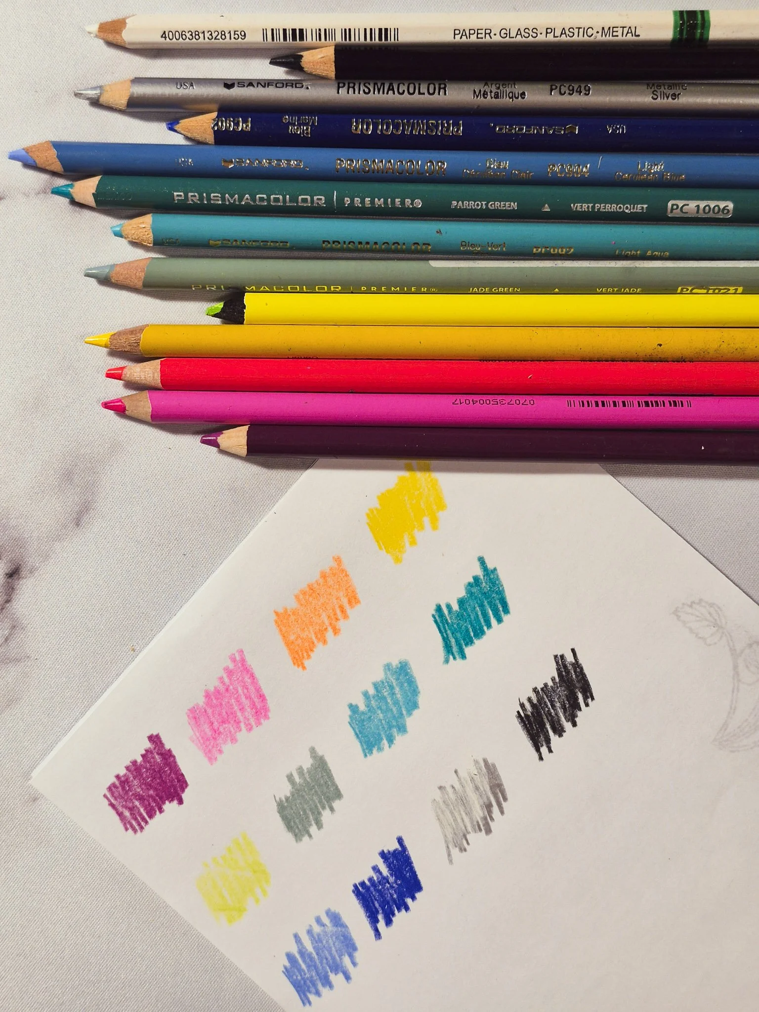 a curated collection of colored pencils inspired by Oliver Jeffers arranges in rainbow order and a scrap paper next to them with the tester swatches scribbled on it.