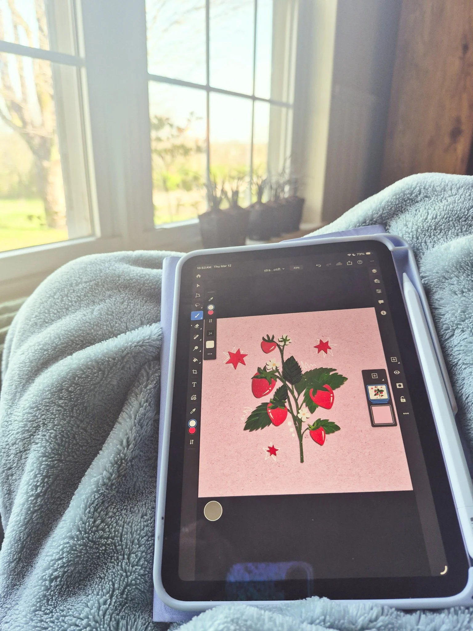 cozy image of an ipad with a strawberry illustration on illustrator's blanket covered lap with a window in the background looking out on a bucolic spring scene