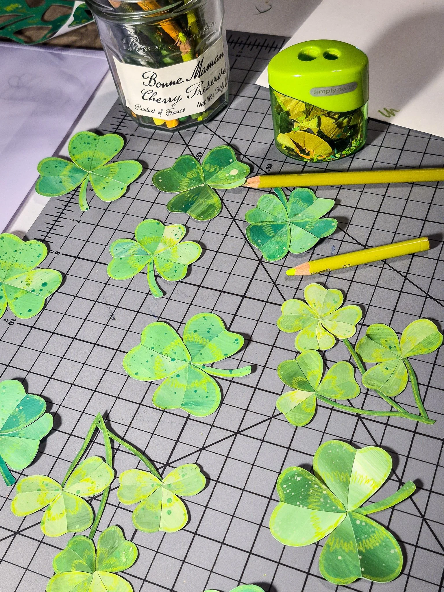 Hand-painted green clover paper cutouts arranged on a grid cutting mat with pencils and art tools.