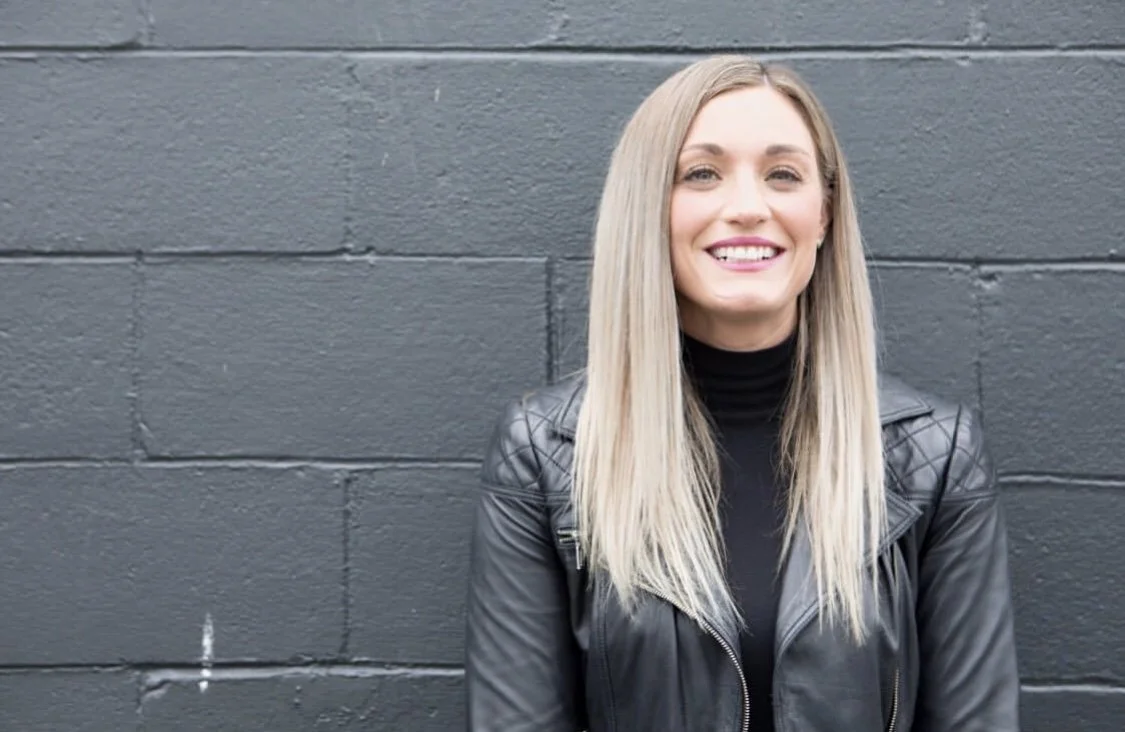 Young woman with long blonde hair, smiling, wearing a black leather jacket and a black turtleneck, standing against a gray brick wall.
