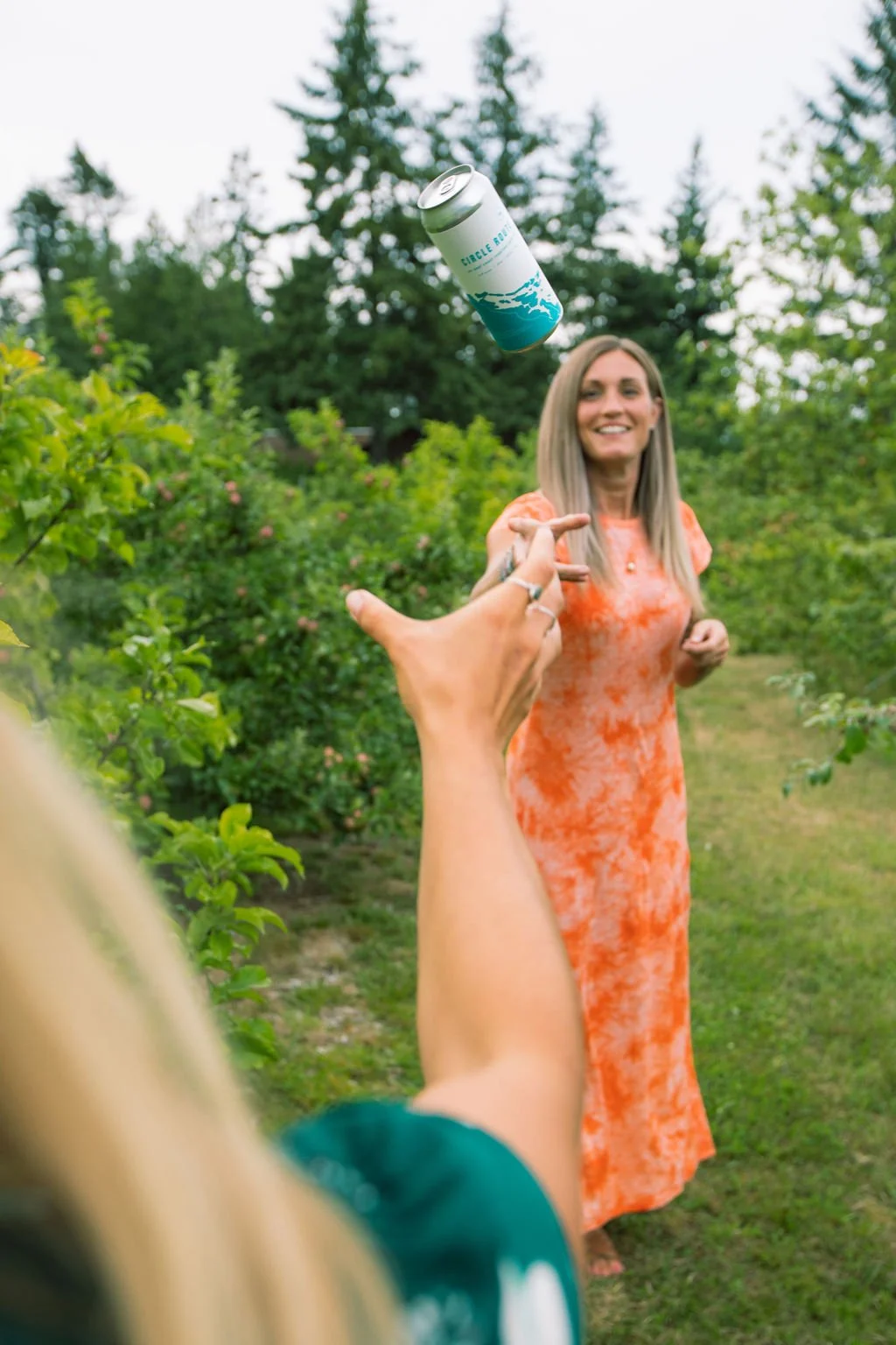 A person throws a can of sparkling water toward a woman standing in a garden, who is smiling in an orange dress.