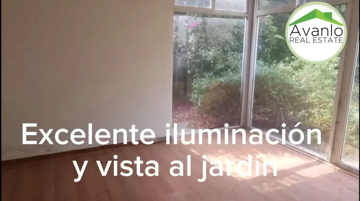 Interior de una habitación con ventanas grandes que dan vista a un jardín, piso de madera, anuncio de bienes raíces con logo de Avanlo y texto que dice 'Excelente iluminación y vista al jardín'.