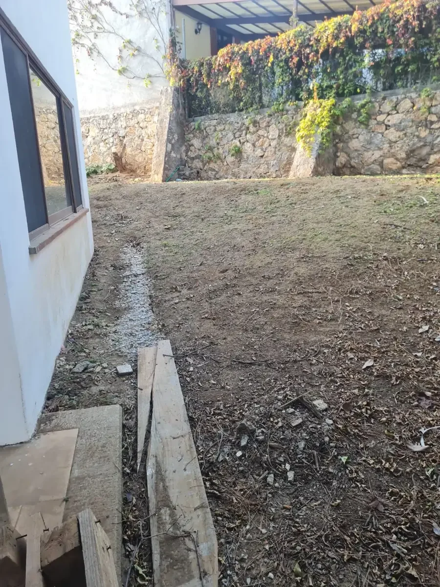 Espacio de jardín en construcción con tierra sin sembrar, bordes de madera y bloques, junto a una pared blanca con ventanas y un muro de piedras con vegetación.