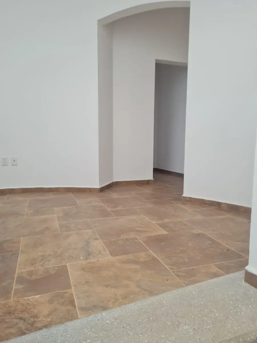 Imagen de una habitación con paredes blancas, suelo de baldosas marrones y un arco que conduce a otro espacio.