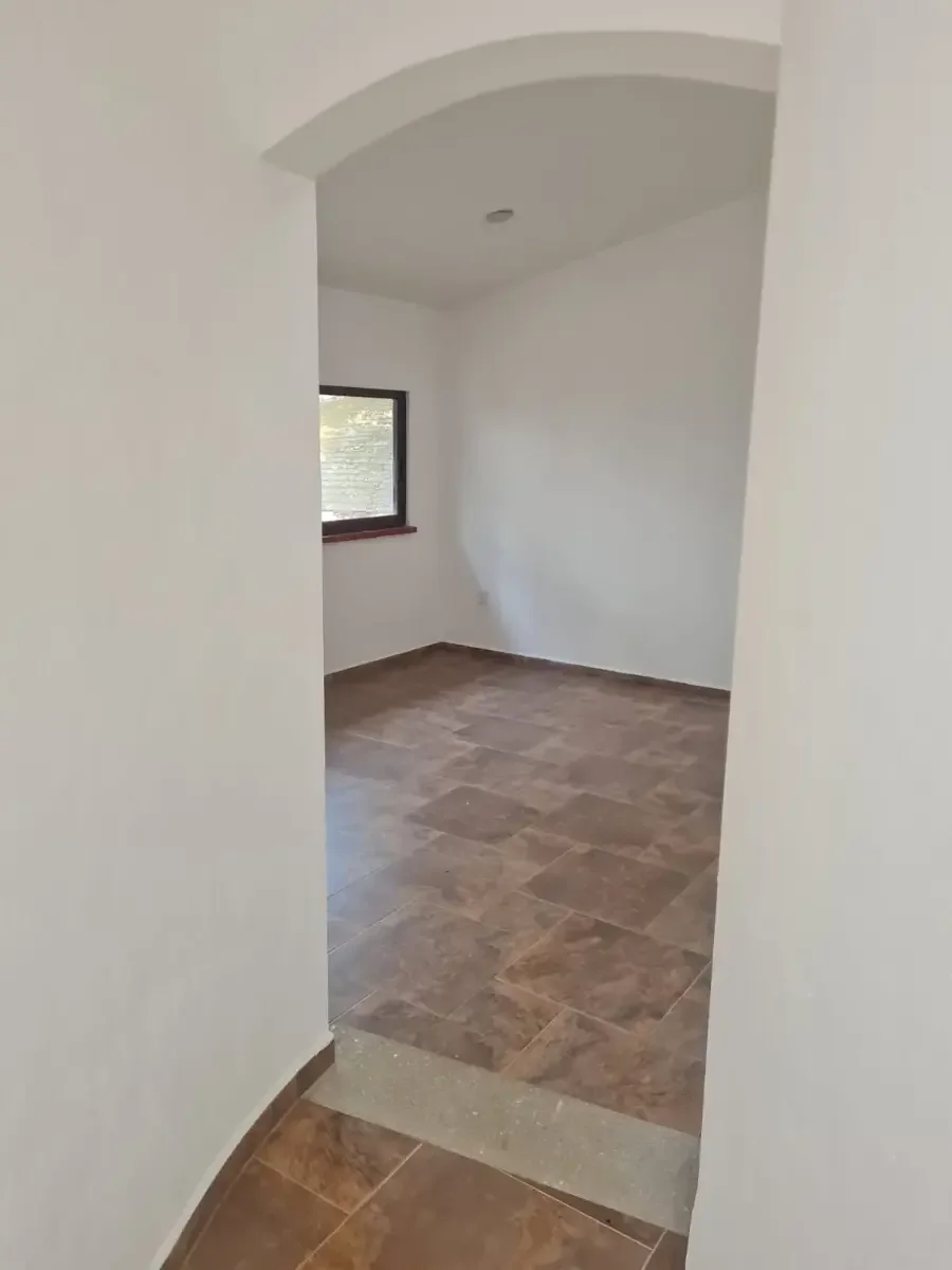 Vista de una habitación vacía con paredes blancas, un ventanal y piso de cerámica marrón.