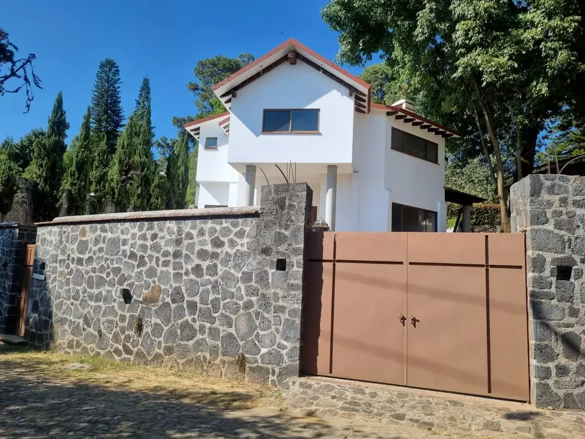 Casa moderna blanca con muro de piedra y portón de metal en un barrio con árboles y cielo despejado.