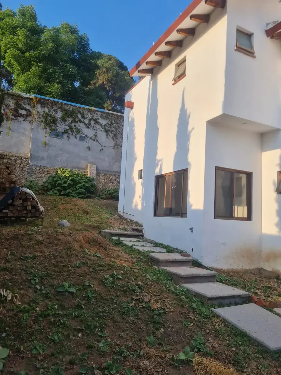 Casa de color blanco vista desde el jardín, con ventanas de madera, sombra de árboles en la pared, y un camino de lajas hacia la entrada.