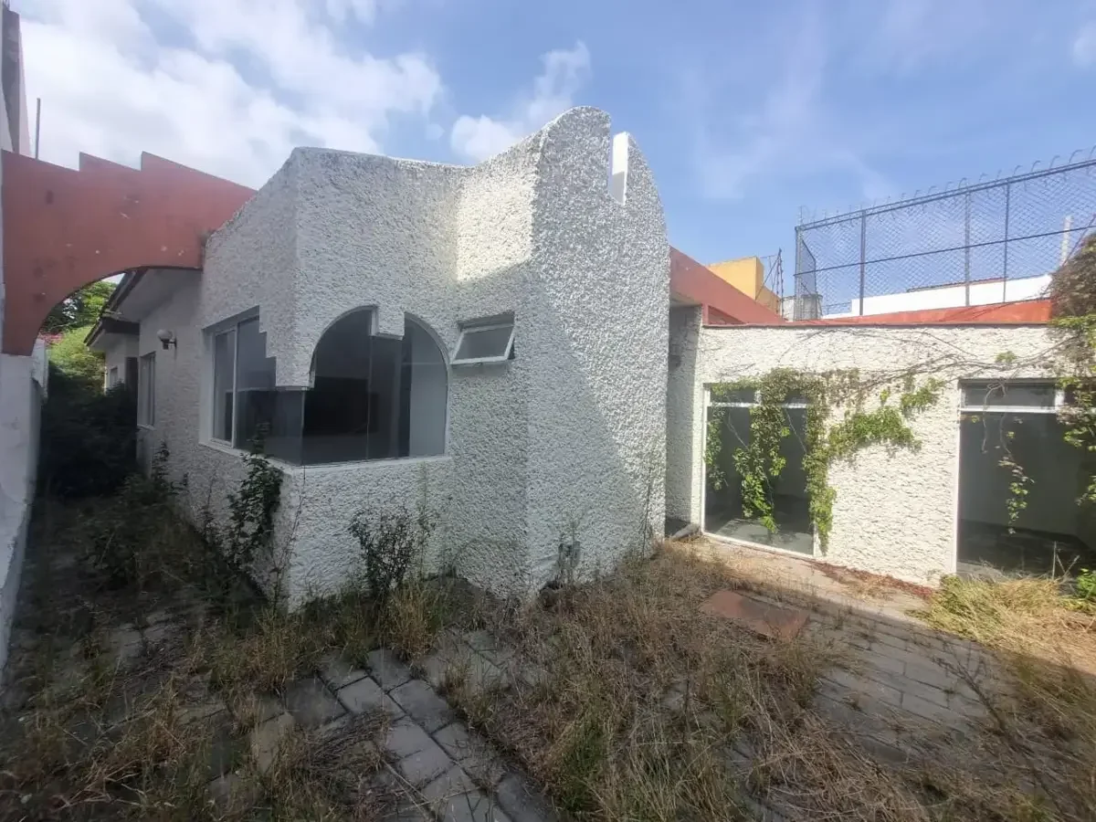 Casa de color blanco con paredes de textura rugosa y ventanas grandes, rodeada de plantas y maleza, con un cielo azul despejado