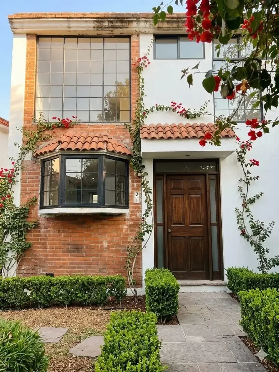 Fachada de casa con pared de ladrillo y paredes blancas, ventanas grandes con marco negro, puerta de madera, techo de tejas rojas, arbustos y plantas en el jardín.