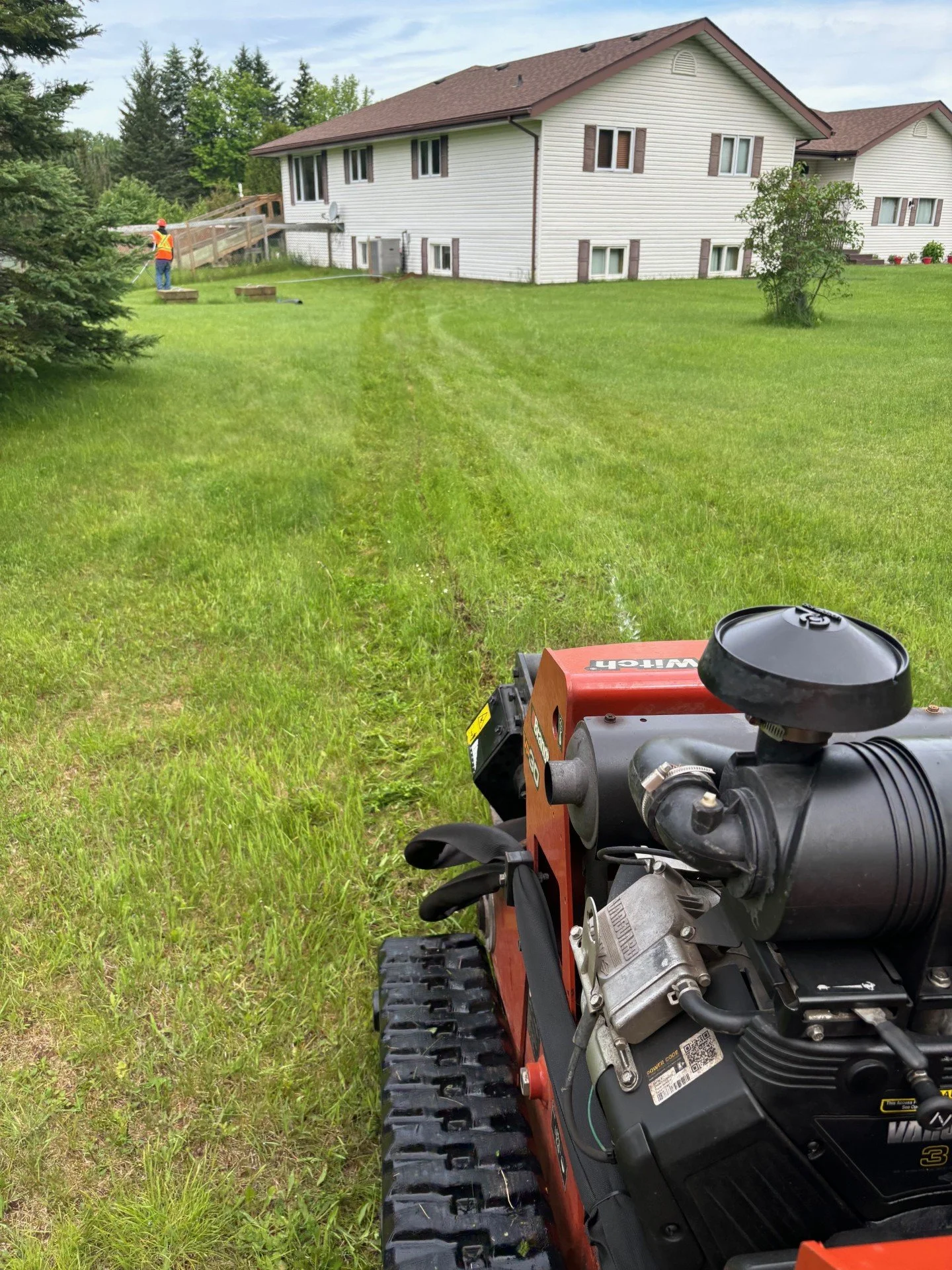 A yard with a lawn mower in the foreground and a person in an orange safety vest working near a wooden deck attached to a house in the background. The house is a two-story white building with multiple windows, surrounded by green grass and trees.