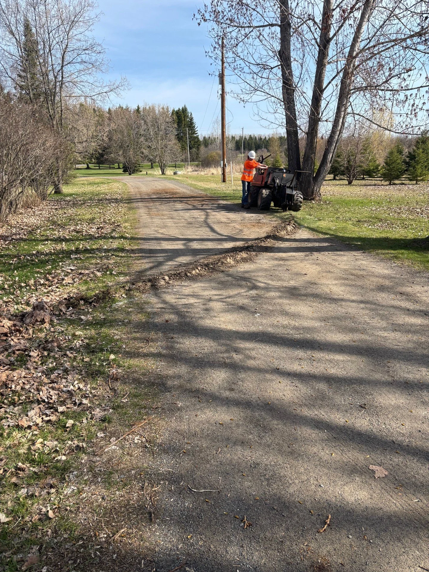 A worker wearing an orange safety vest and white helmet operates a piece of heavy equipment near a large tree on a dirt and gravel rural road under a partly cloudy sky.
