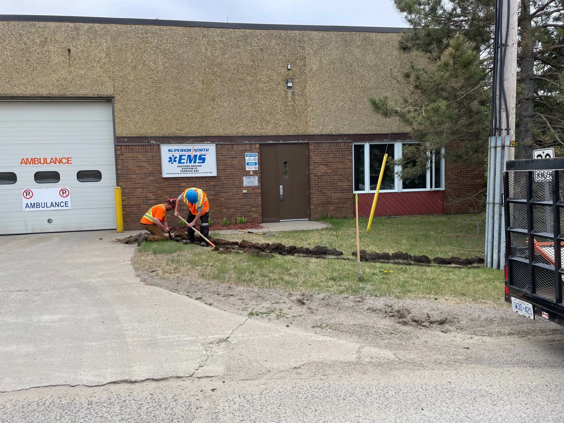 Two construction workers in safety vests and helmets working on a trench outside a building marked 'Superior North EMS' and 'Ambulance' garage, with a grassy area and utility pole nearby.