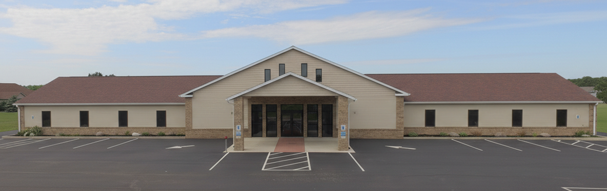 A single-story building with beige siding, brick accents, and a shingle roof, featuring a central entrance with glass doors and windows, surrounded by an asphalt parking lot with marked spaces.